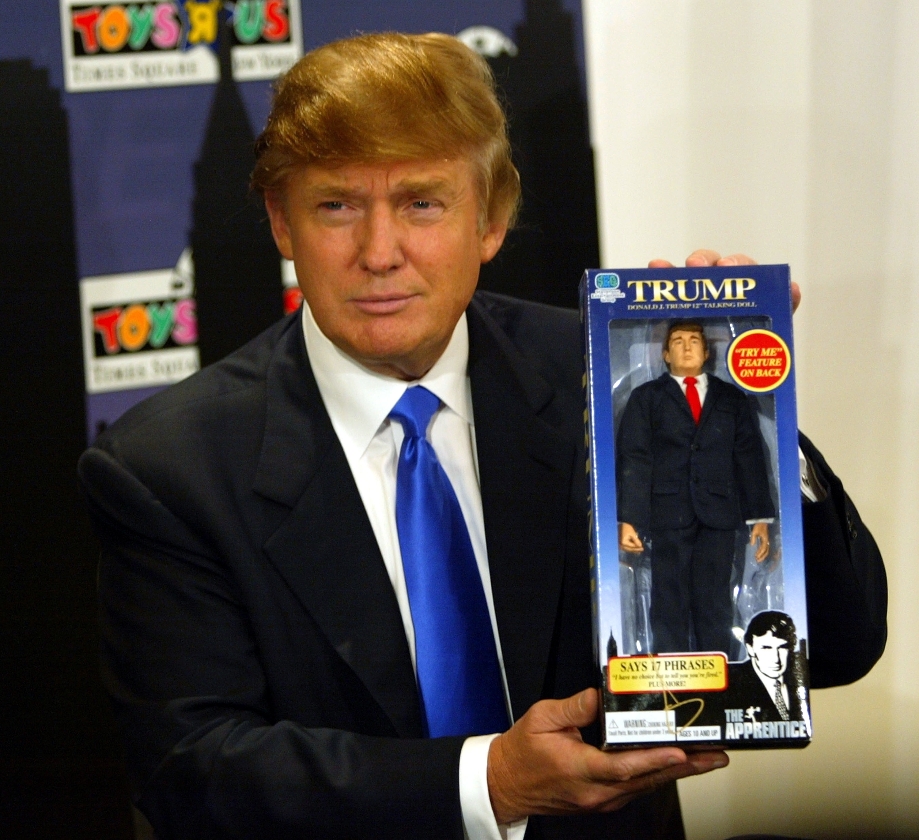 Trump holding a Trump action figure in a box labeled "The Apprentice" at a Toys R Us event