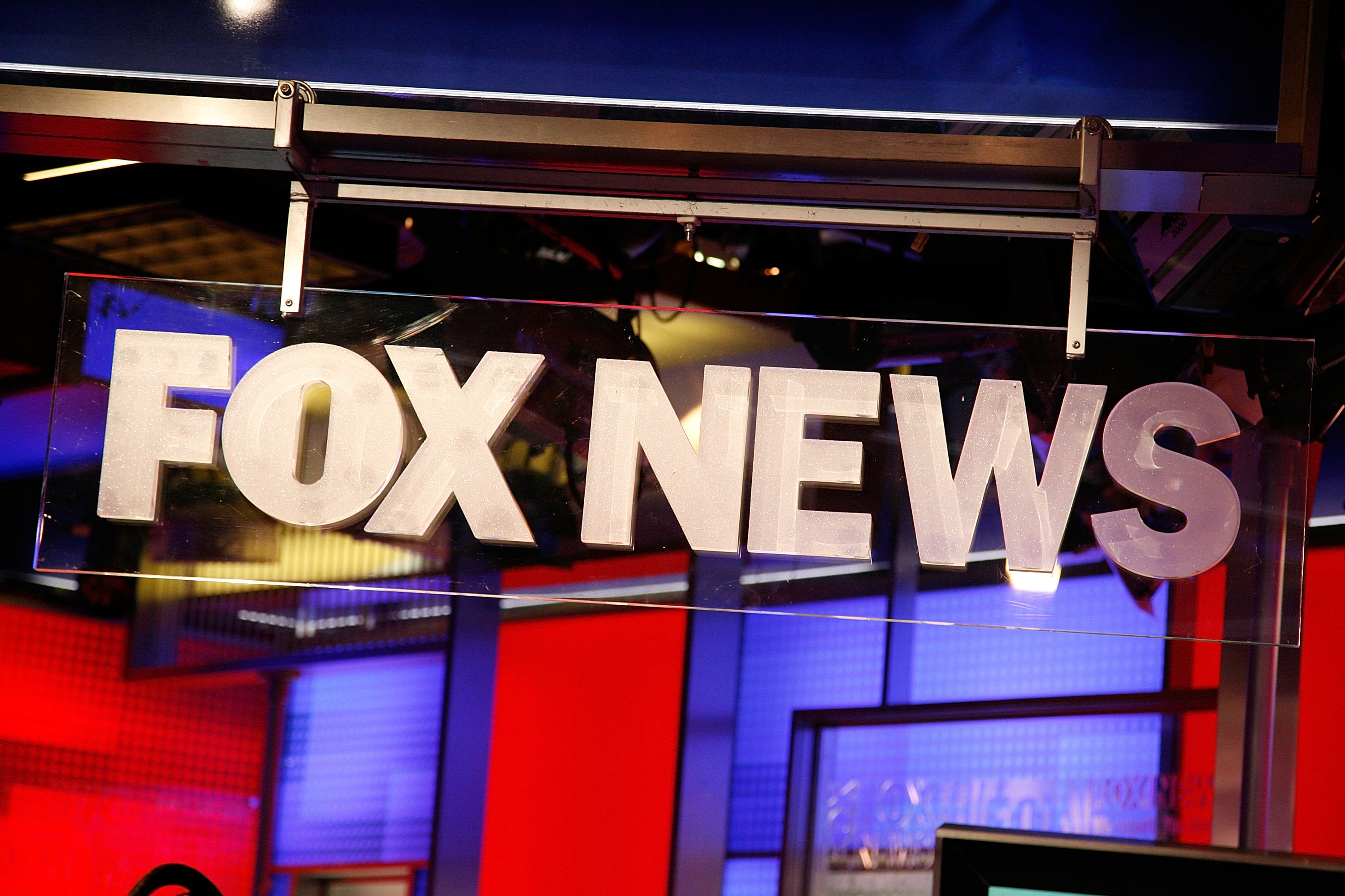 Sign displaying "FOX NEWS" inside a broadcast studio with visible blue and red lit walls in the background
