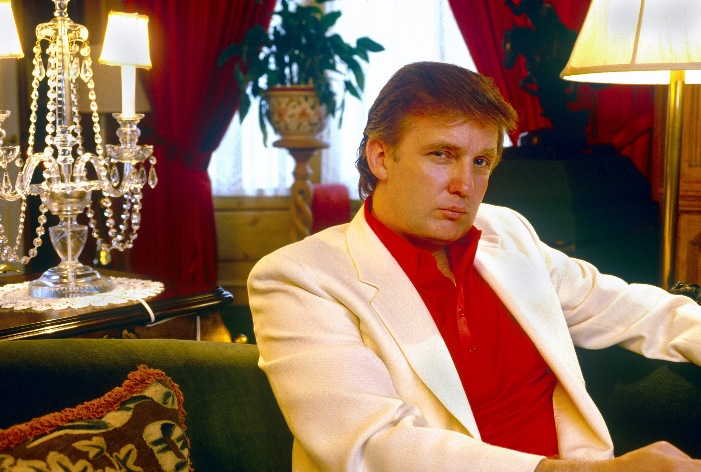 Trump sits in a lavish room with ornate decor and a chandelier, wearing a white suit jacket over a red shirt