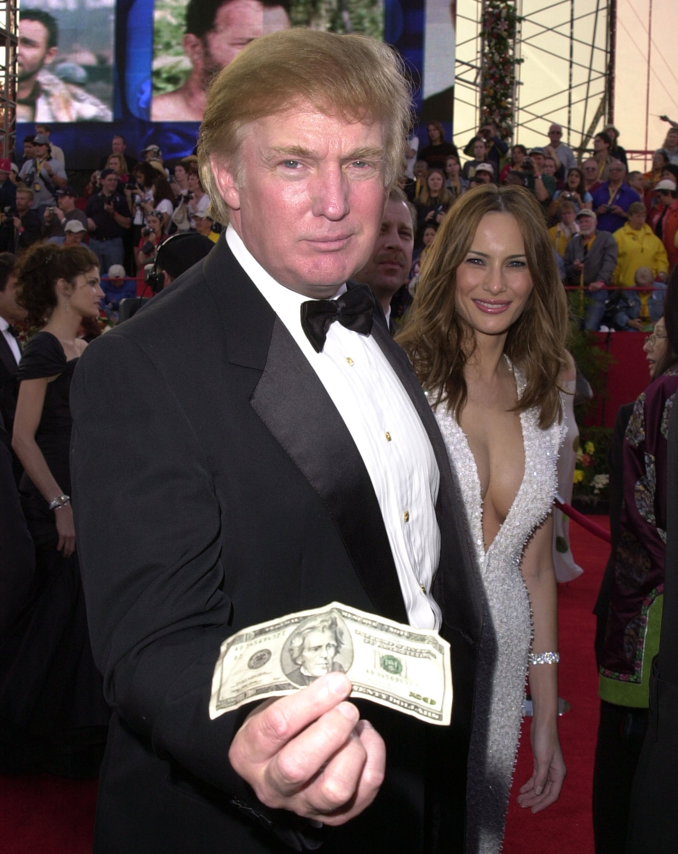 Donald Trump in a tuxedo holds a banknote on a red carpet, with a woman in a low-cut, embellished gown in the background