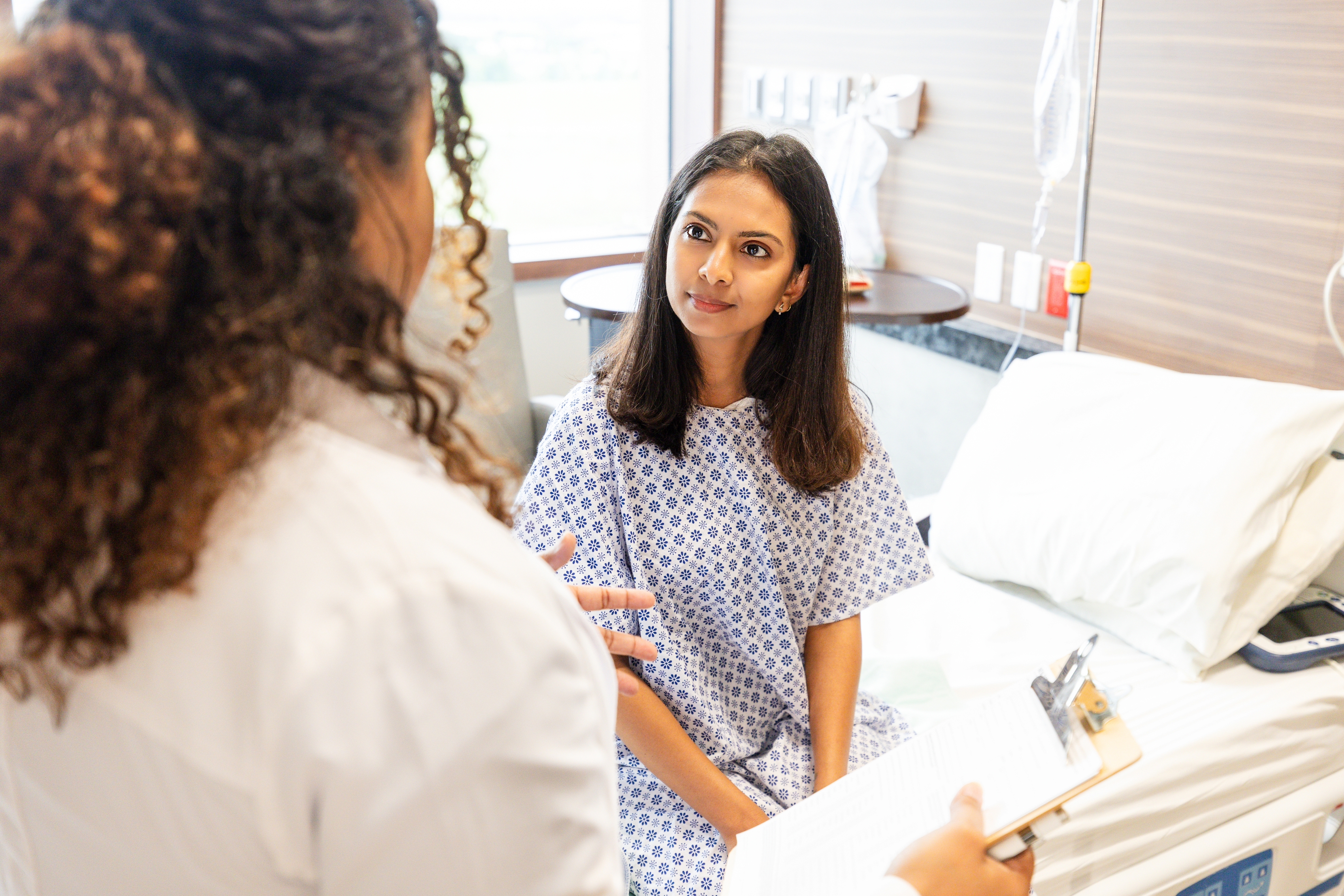 En pasient i en sykehuskjole lytter oppmerksomt til en helsepersonell som holder et utklippstavle, i et sykehusrom