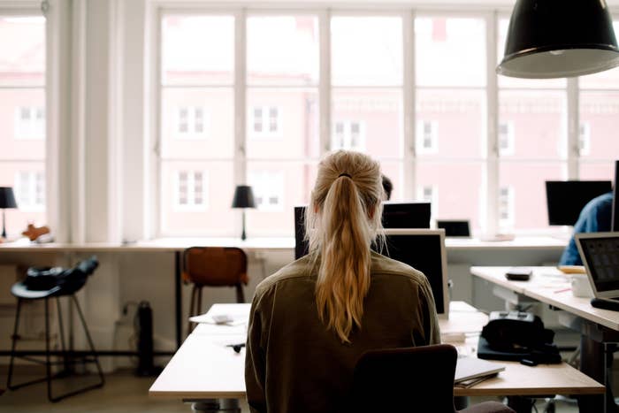 Person with long hair sits at a desk in a backlit office, facing computer screens. Large windows line the back wall, letting in natural light