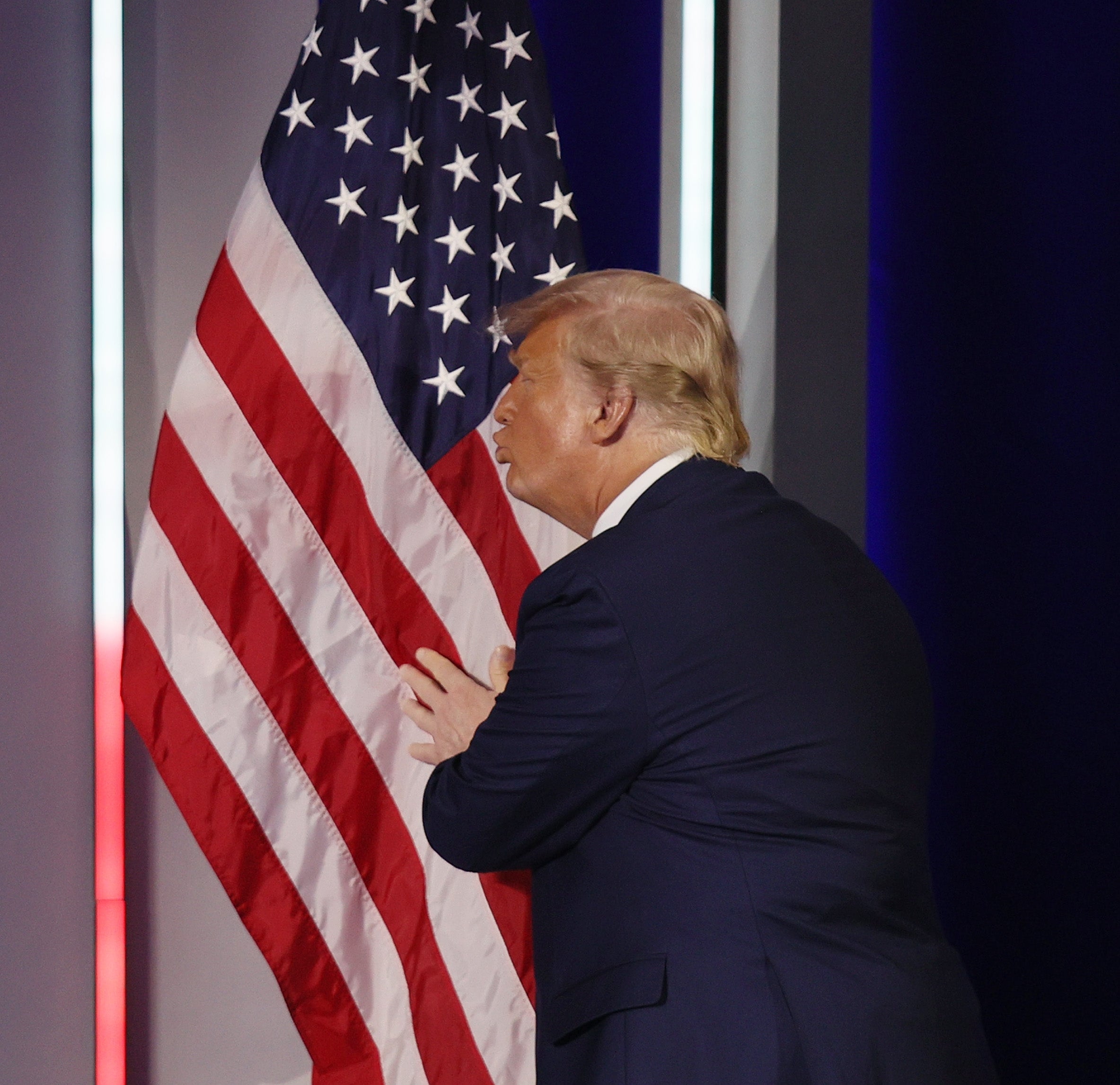 Trump in a suit embraces an American flag on stage, expressing affection towards the flag