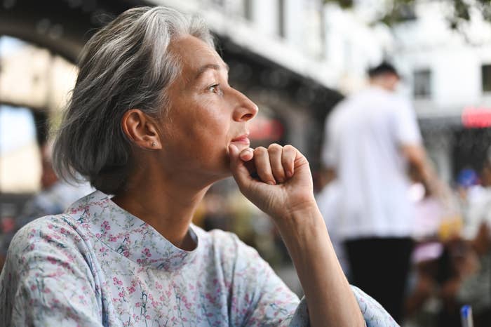 Person with gray hair in floral top sits outdoors, looking contemplative, with blurred people in the background