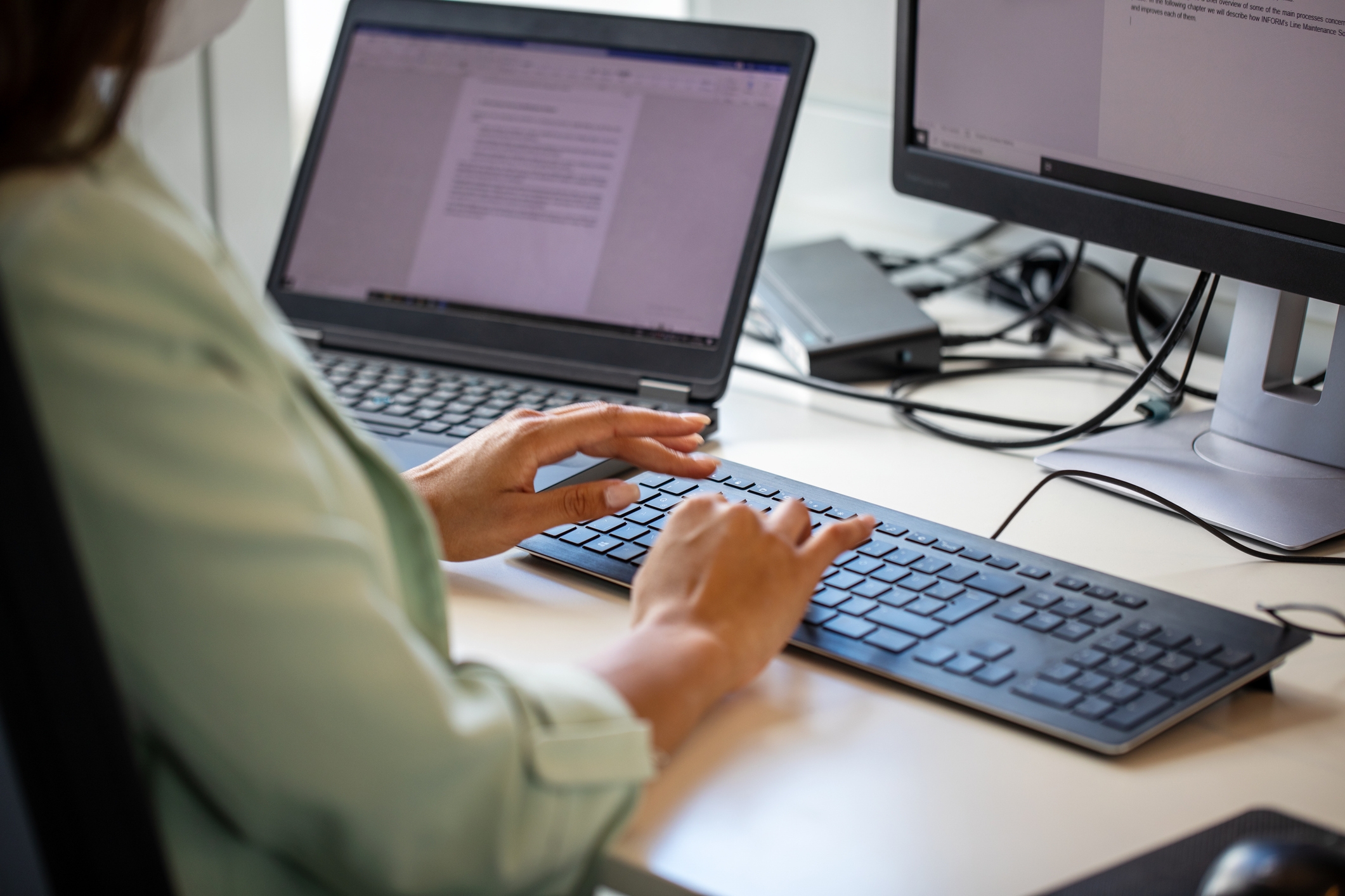 Person typing on a keyboard at a desk with two monitors showing documents