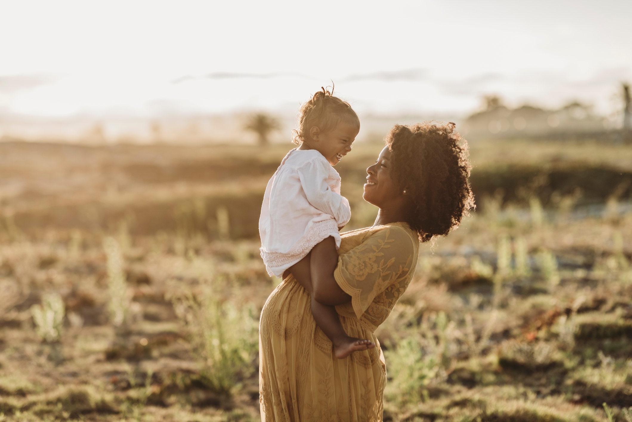 A woman in a flowing dress holds a child, sharing a joyful moment together in an open field, with warm sunlight in the background