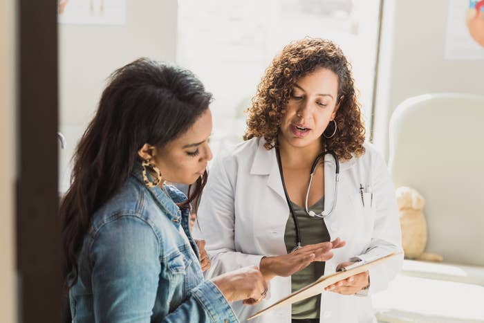 Two women in discussion; one with a lab coat and stethoscope gestures to a tablet held by the other, possibly discussing medical information