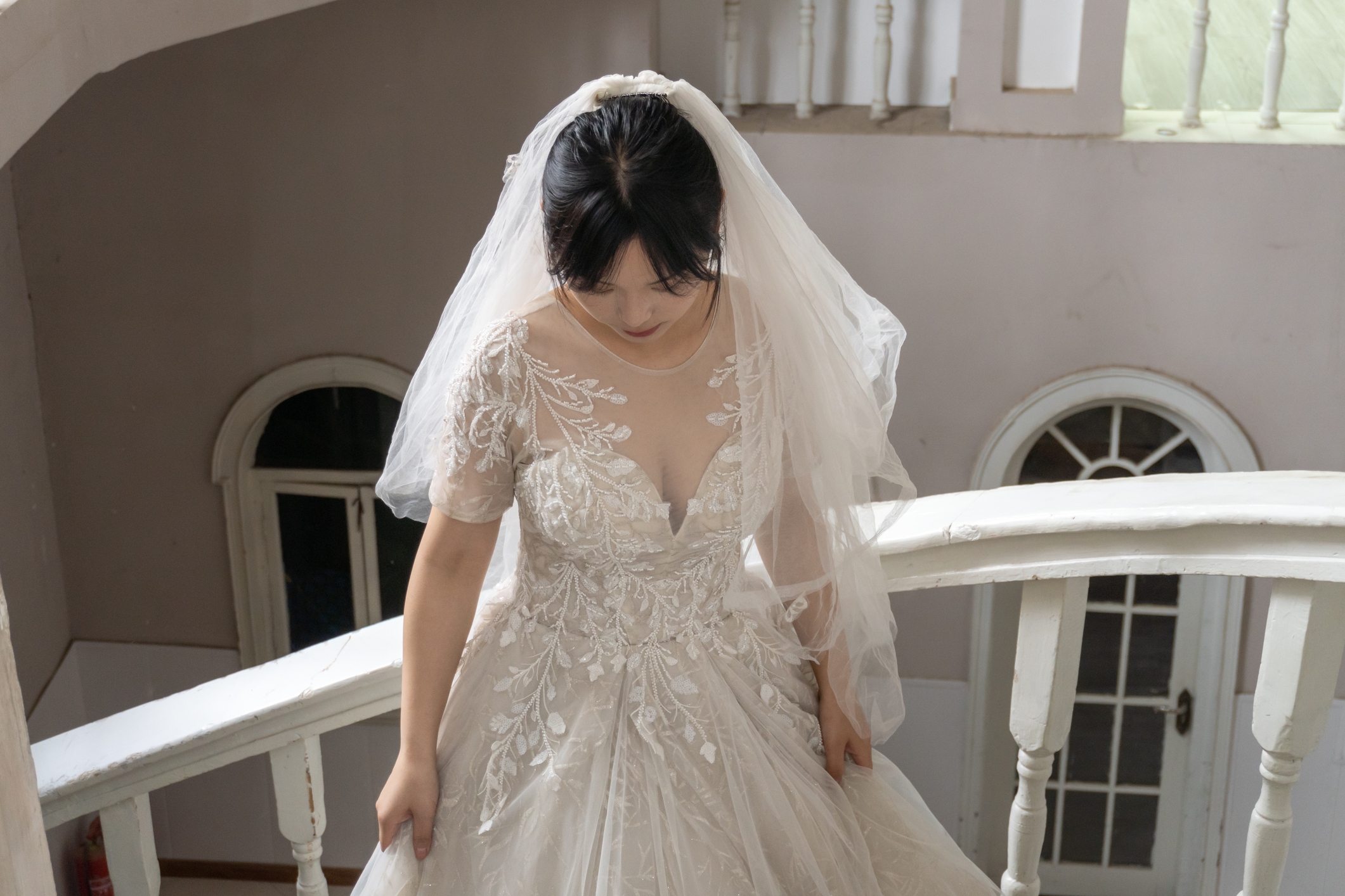 Bride in a lace wedding dress with a floral pattern and veil, standing on a staircase and looking down
