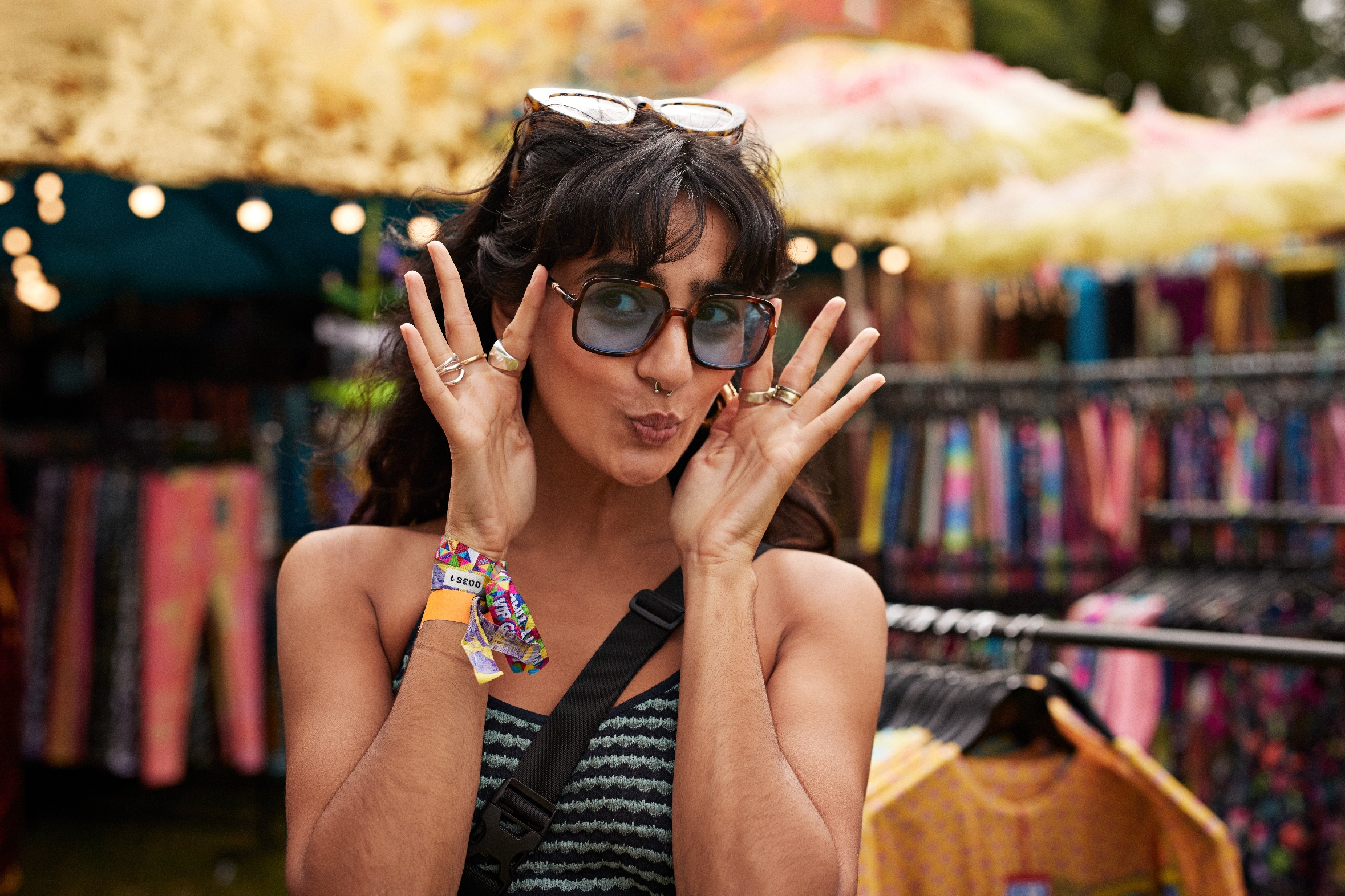 Person playfully posing with peace signs near their face, wearing sunglasses and colorful wristbands, in a vibrant outdoor market setting