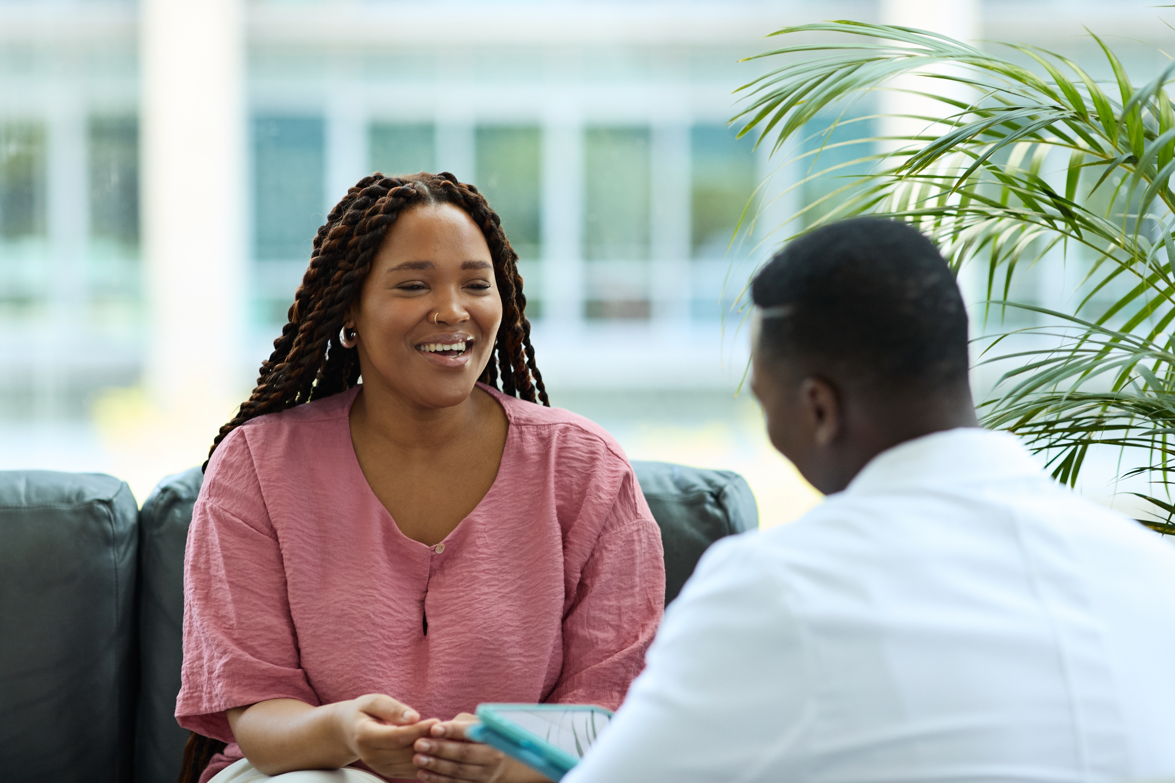 Two people sit on a couch engaged in conversation. The person on the left smiles warmly, wearing a textured shirt. A plant is visible nearby