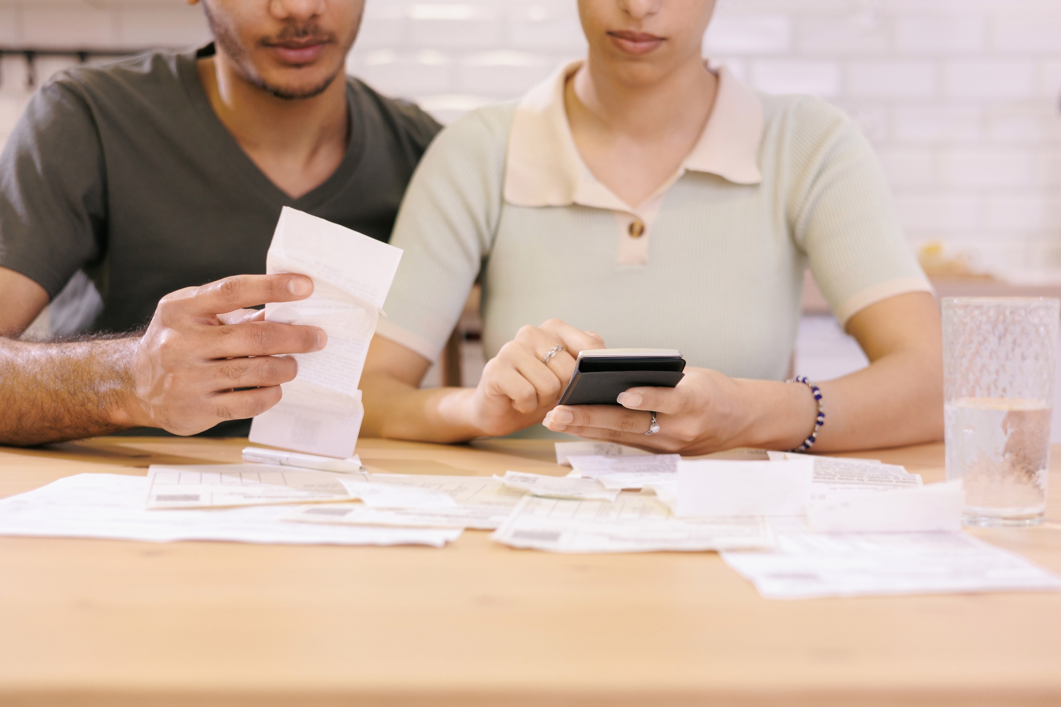 A couple reviews receipts and works on finances at a table; they use a smartphone and calculator, surrounded by bills and documents