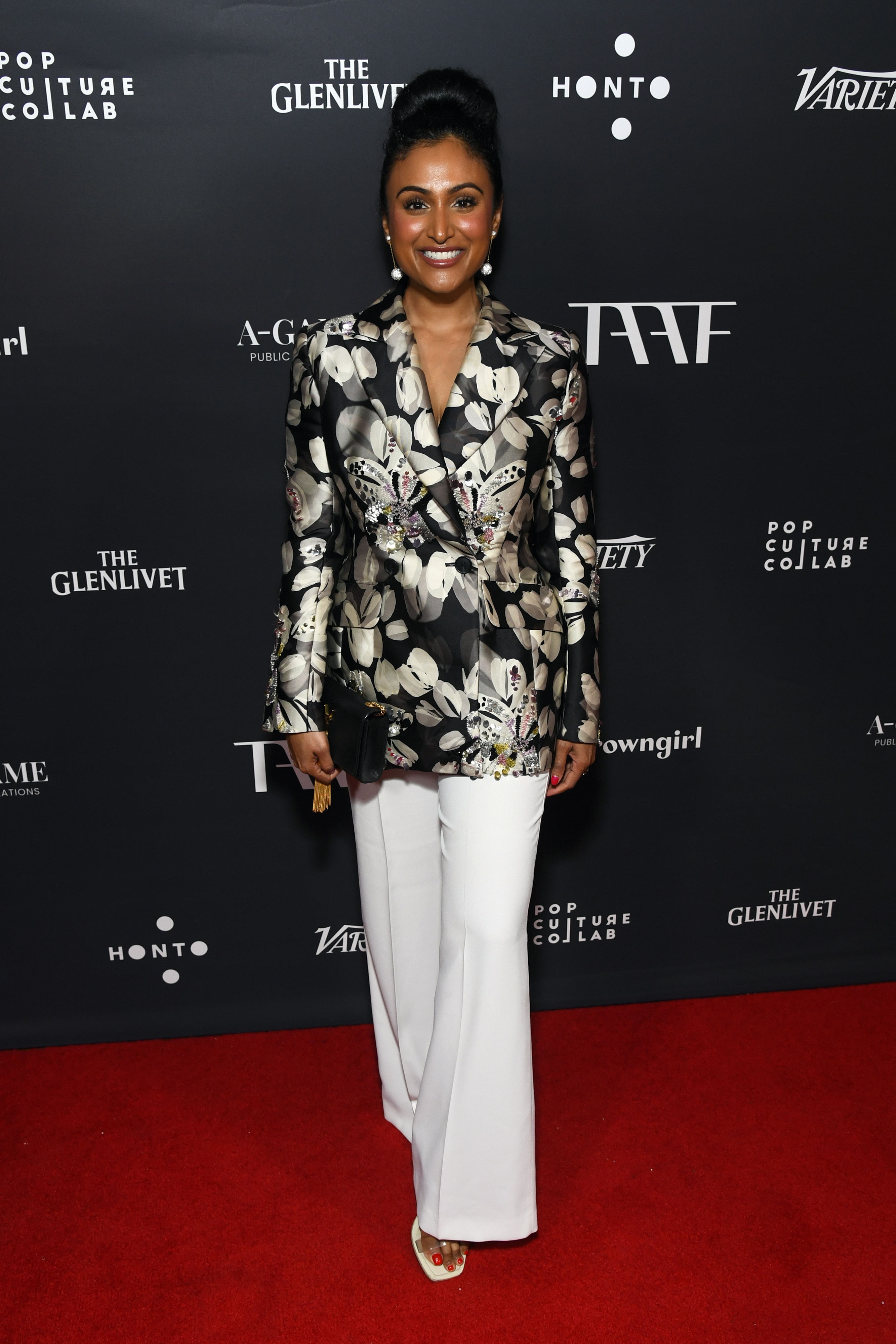 Person på rød teppe i en blomstermønstret blazer og hvite bukser, smilende mens du holder en clutch. Hendelseslogoer i bakgrunnen