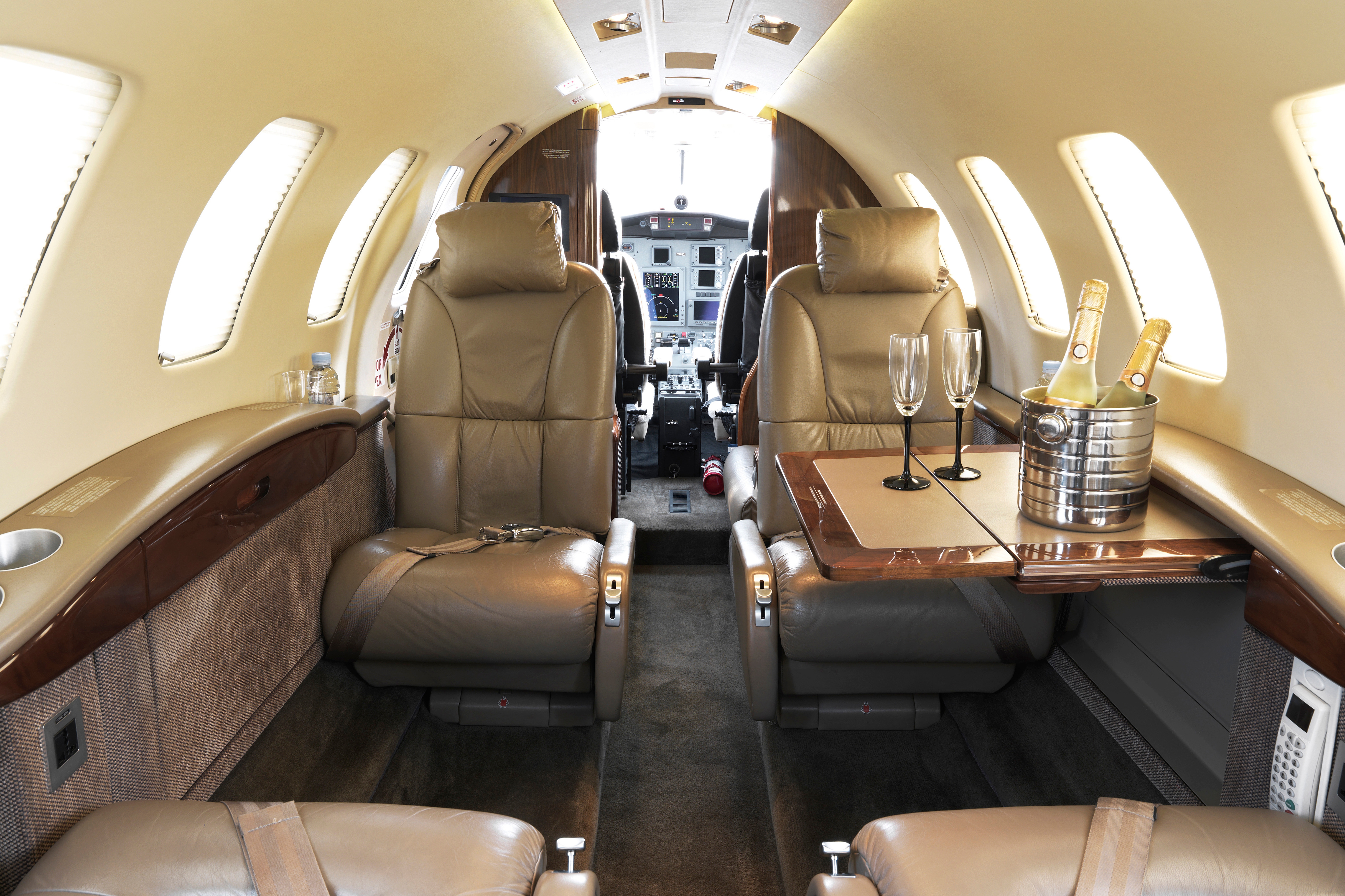 Luxury private jet interior with leather seats, two tables, champagne bottles, and glasses. Cockpit visible in the background