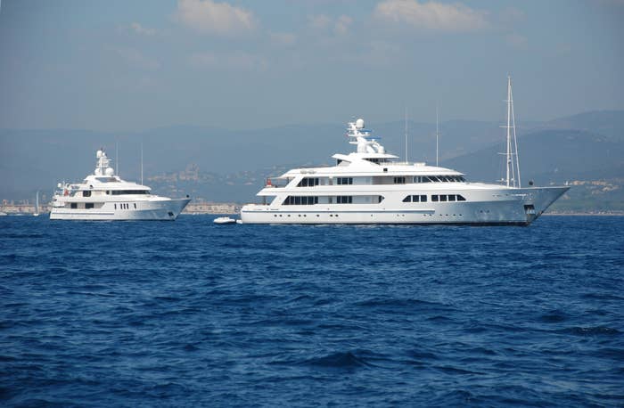 Two large yachts anchored on a calm sea with distant hills in the background under a partly cloudy sky