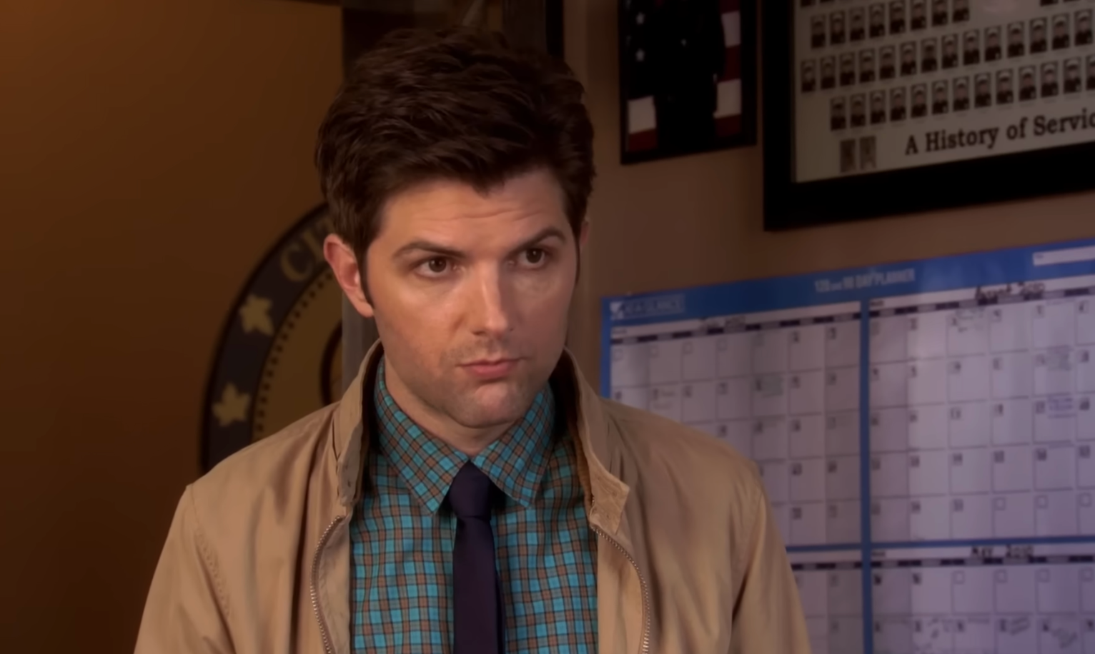 Person in a checked shirt and tie with a neutral expression, standing indoors by a calendar