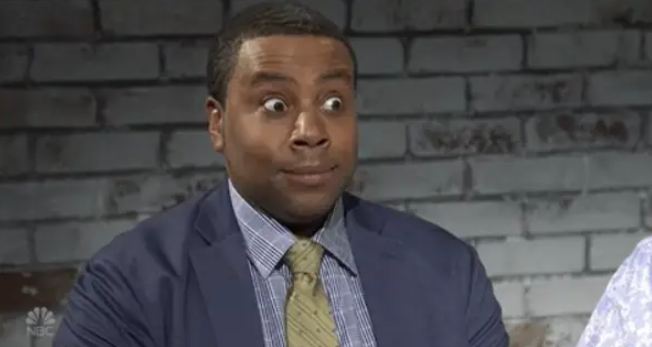 A person with a surprised expression is wearing a suit and tie in front of a brick wall