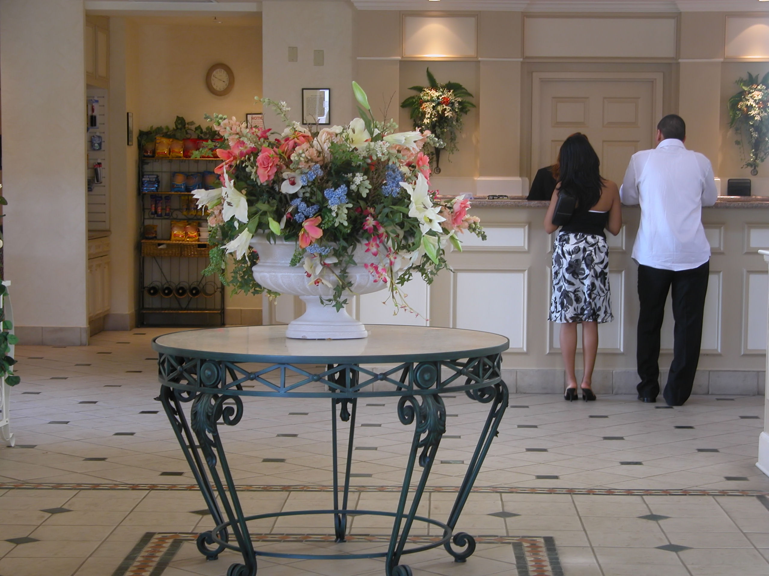 A couple stands at a reception desk in a hotel lobby. A decorative floral arrangement is on a round table in the foreground