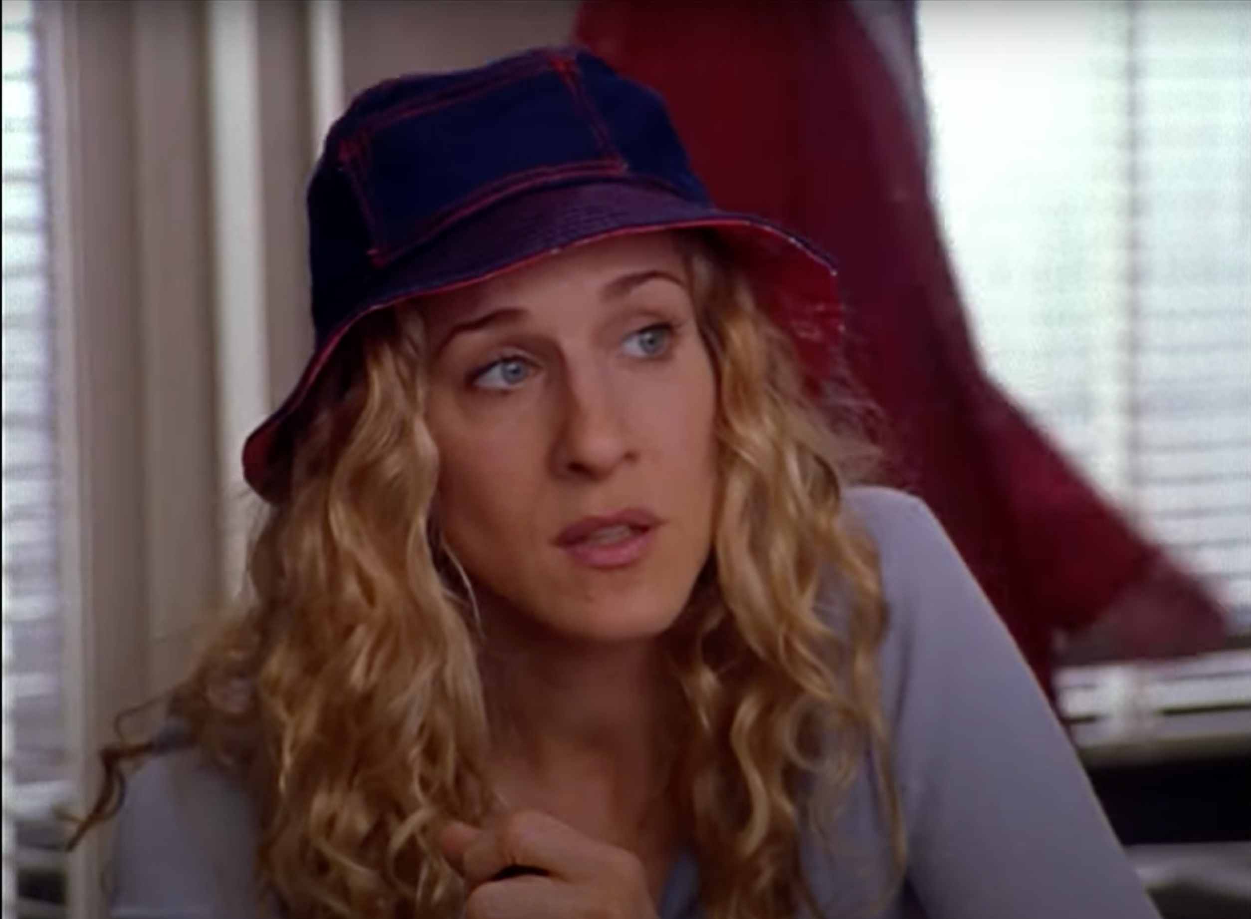 Person with long curly hair wearing a bucket hat and a casual top, sitting indoors, appearing to be in conversation