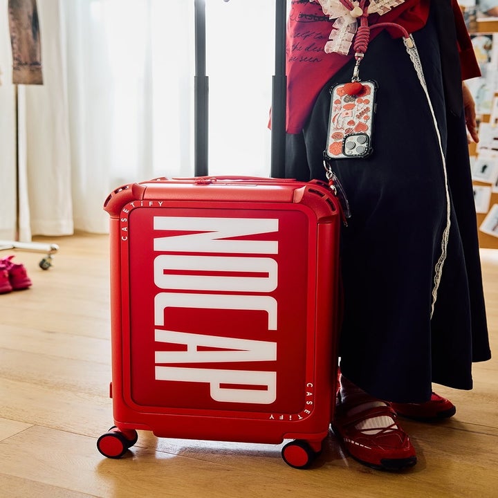 Red suitcase with large white text "NO CAP" on the front, next to a person in stylish clothing and red shoes, standing indoors