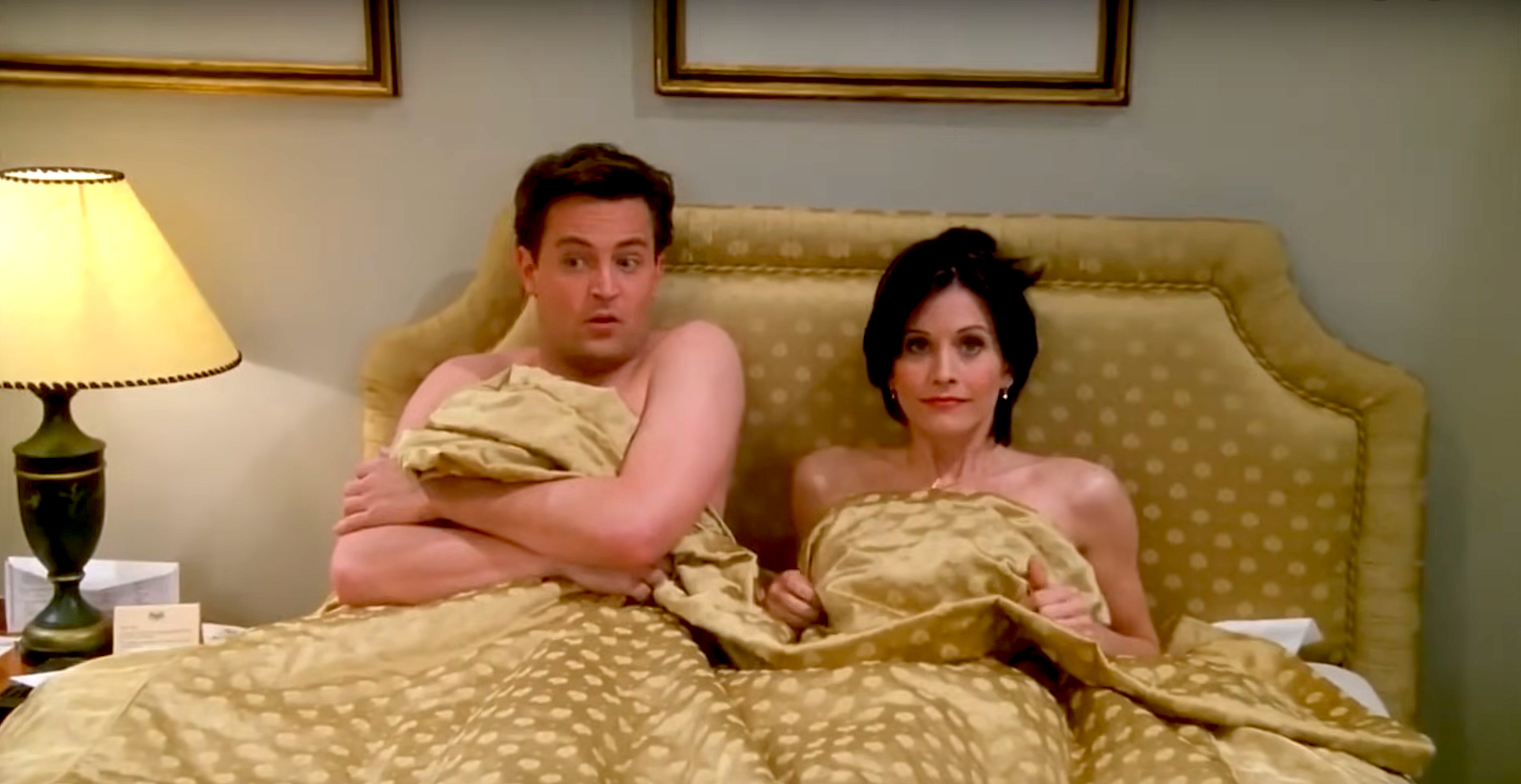 Two people in bed looking surprised, partially covered by a blanket