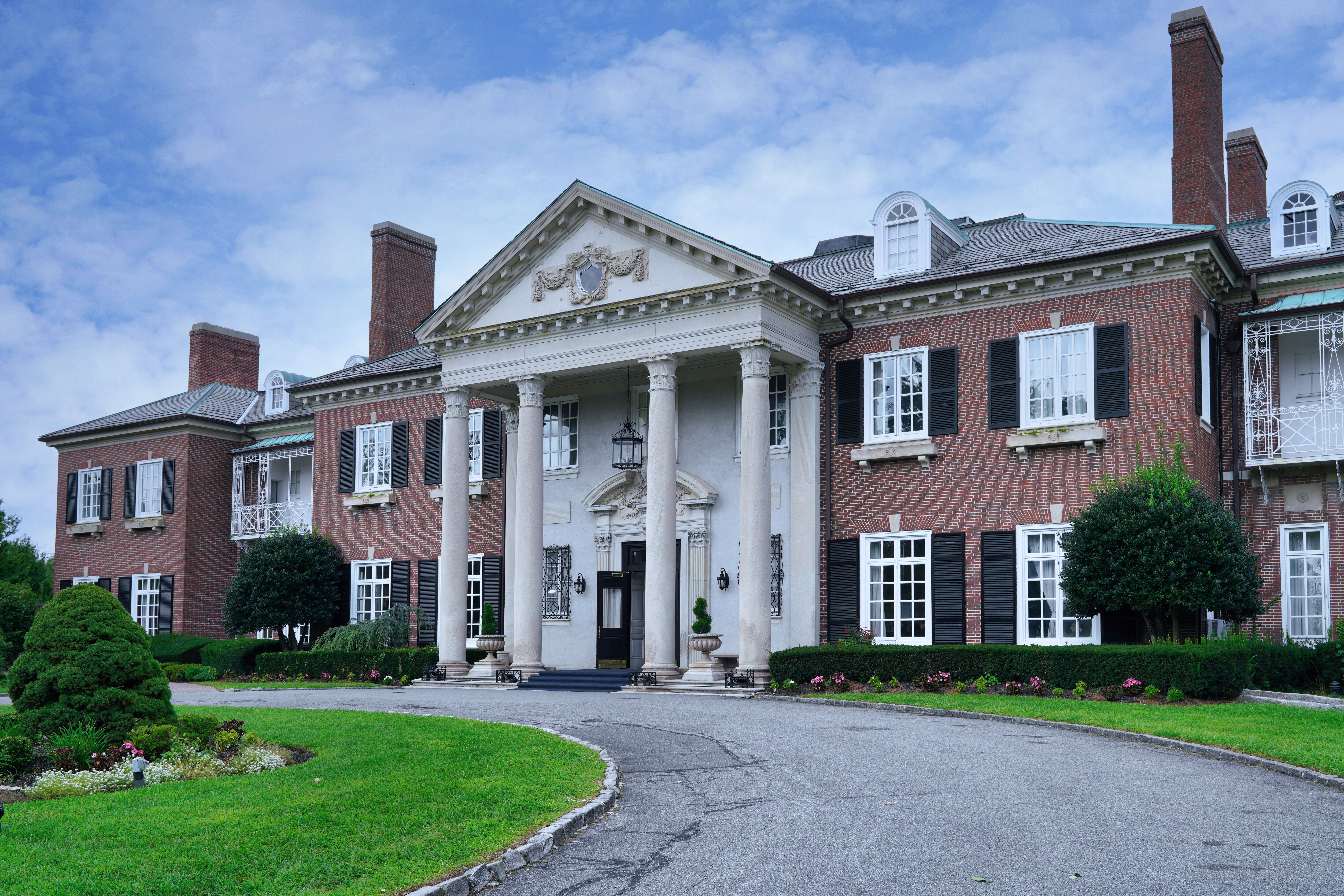 Large colonial-style mansion with tall columns, red brick facade, multiple chimneys, and a circular driveway surrounded by manicured greenery
