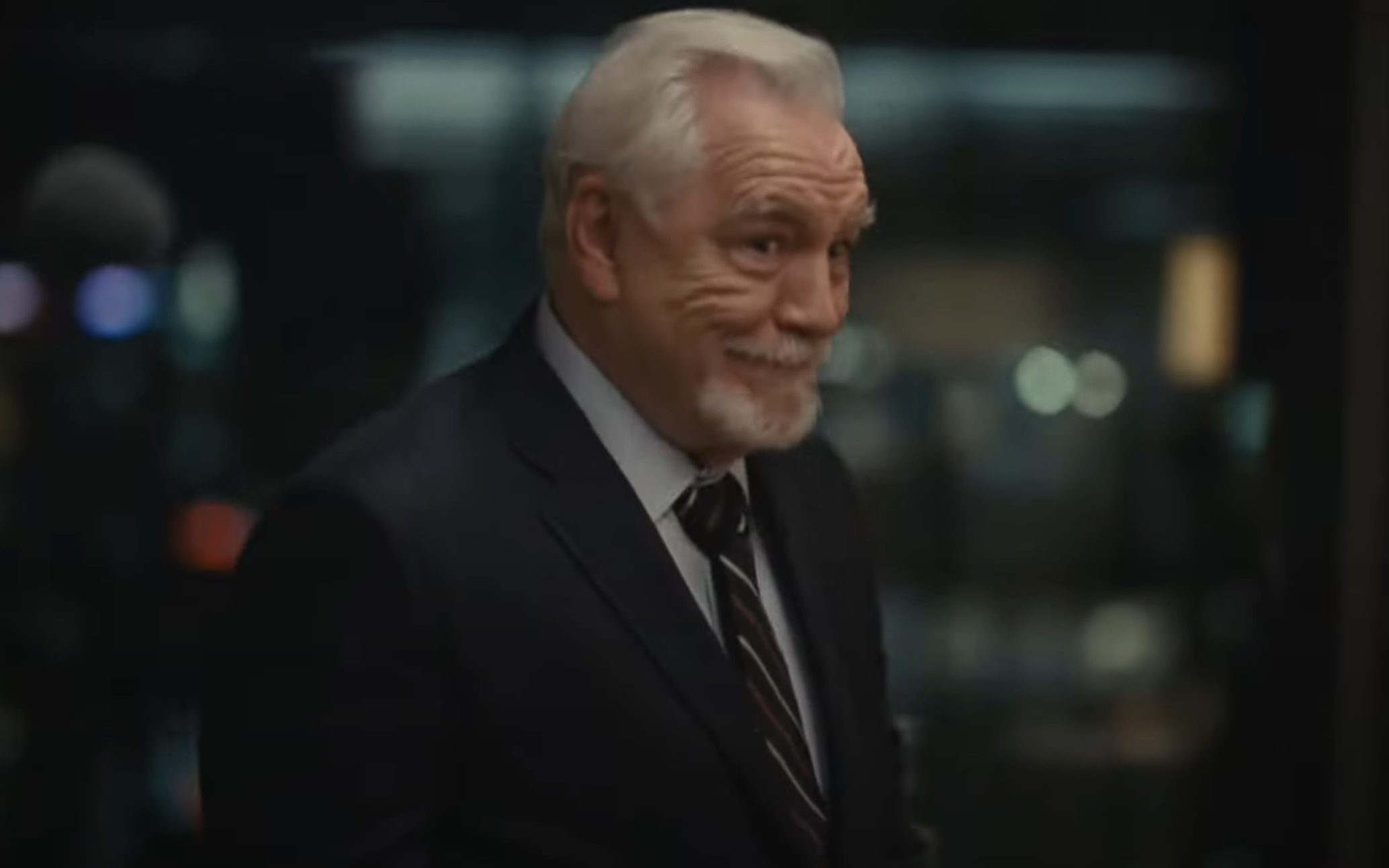 Older man in a suit, smiling slightly, in a dimly lit environment