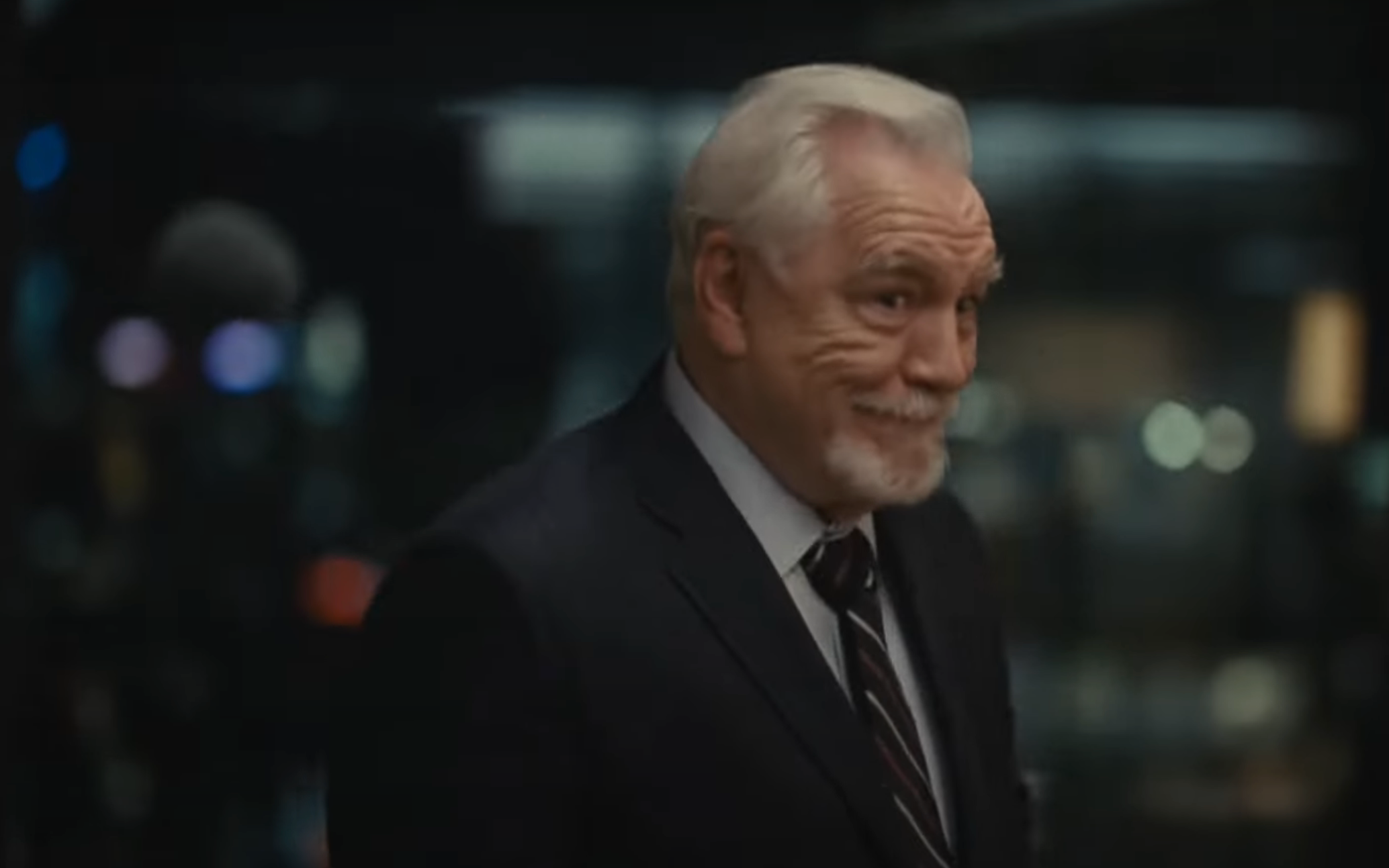 Older man in a suit, smiling slightly, in a dimly lit environment