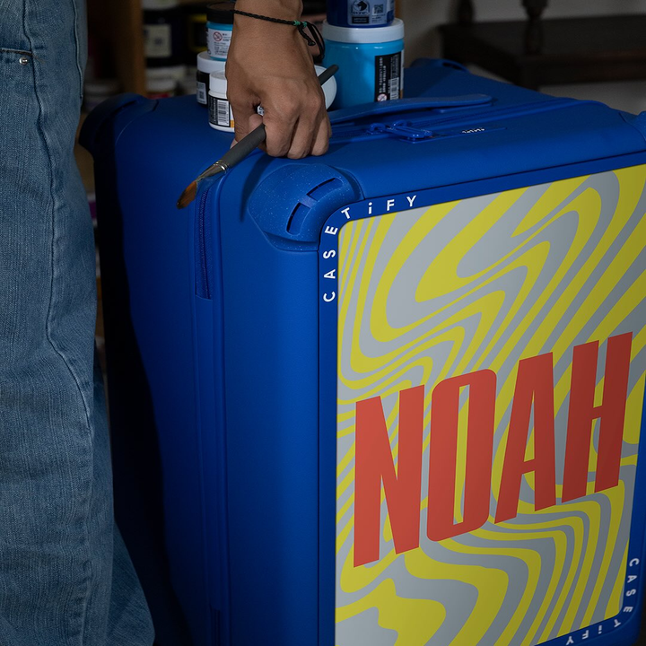 Person holding blue suitcase with a bold design, featuring the brand "CASETiFY" and the name "NOAH" prominently displayed. Denim clothing is visible