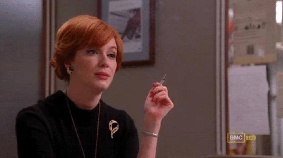 Person in vintage-style clothing seated at a table, holding a pen thoughtfully. Office setting in the background