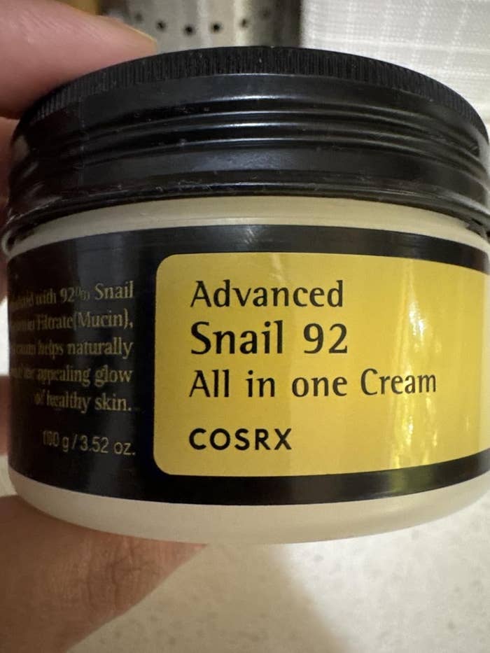 A jar of COSRX Advanced Snail 92 All in one Cream is held in a hand, featuring skincare product information on the label