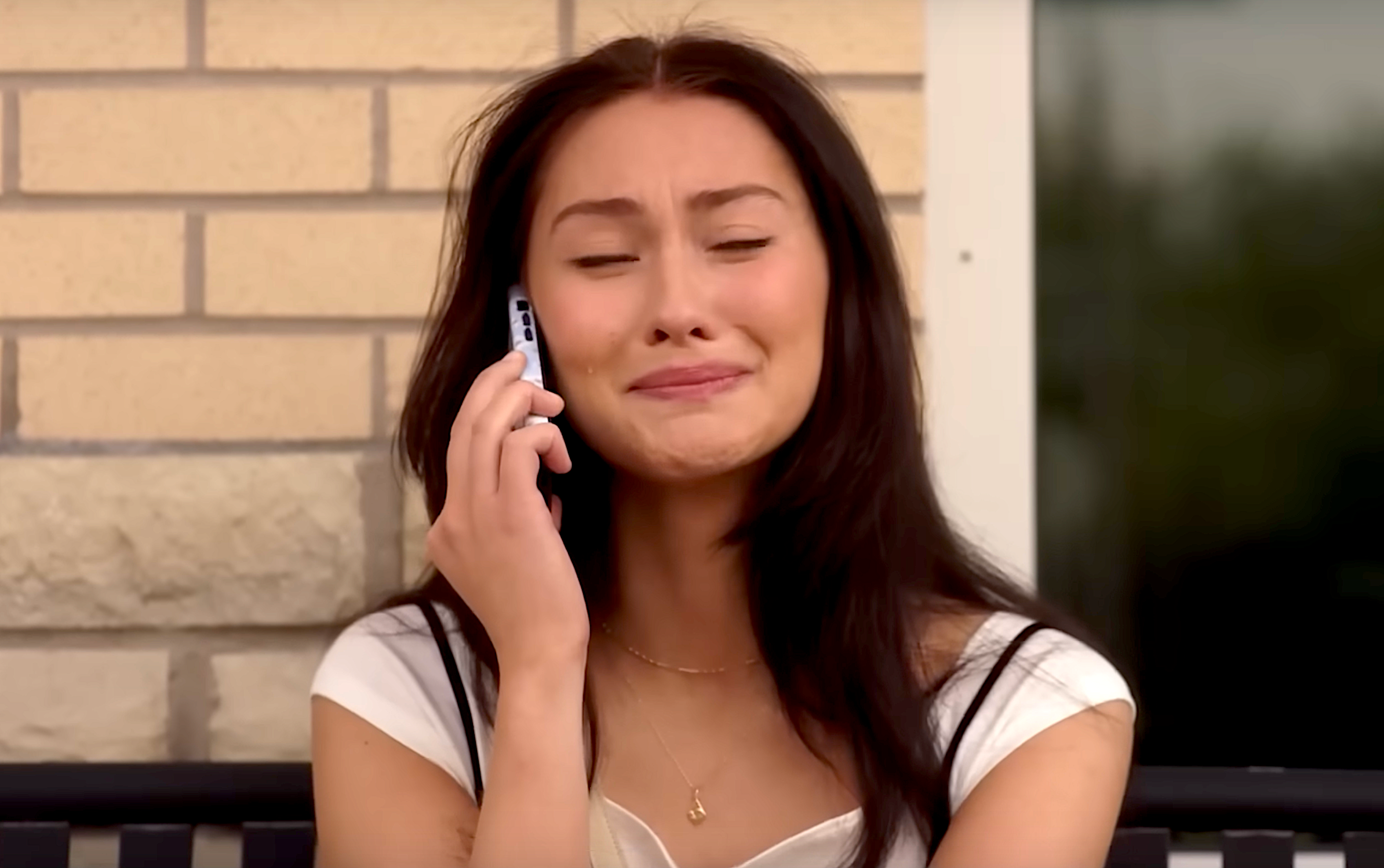 Person sitting outdoors, holding a phone to their ear, appears emotional with eyes closed and a smile