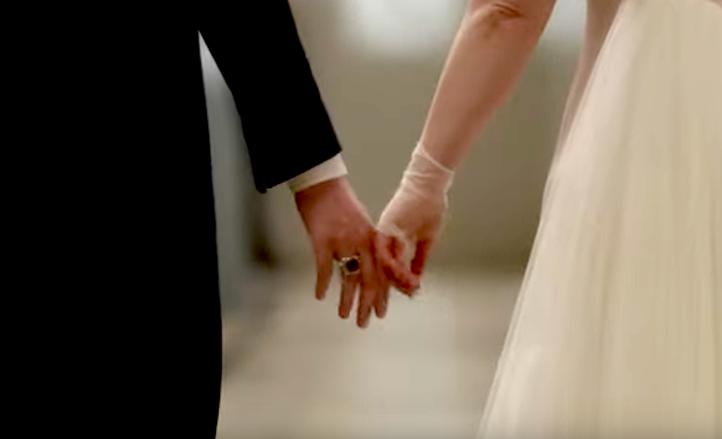 A person in formal attire and another in a wedding dress holding hands, focusing on their joined hands