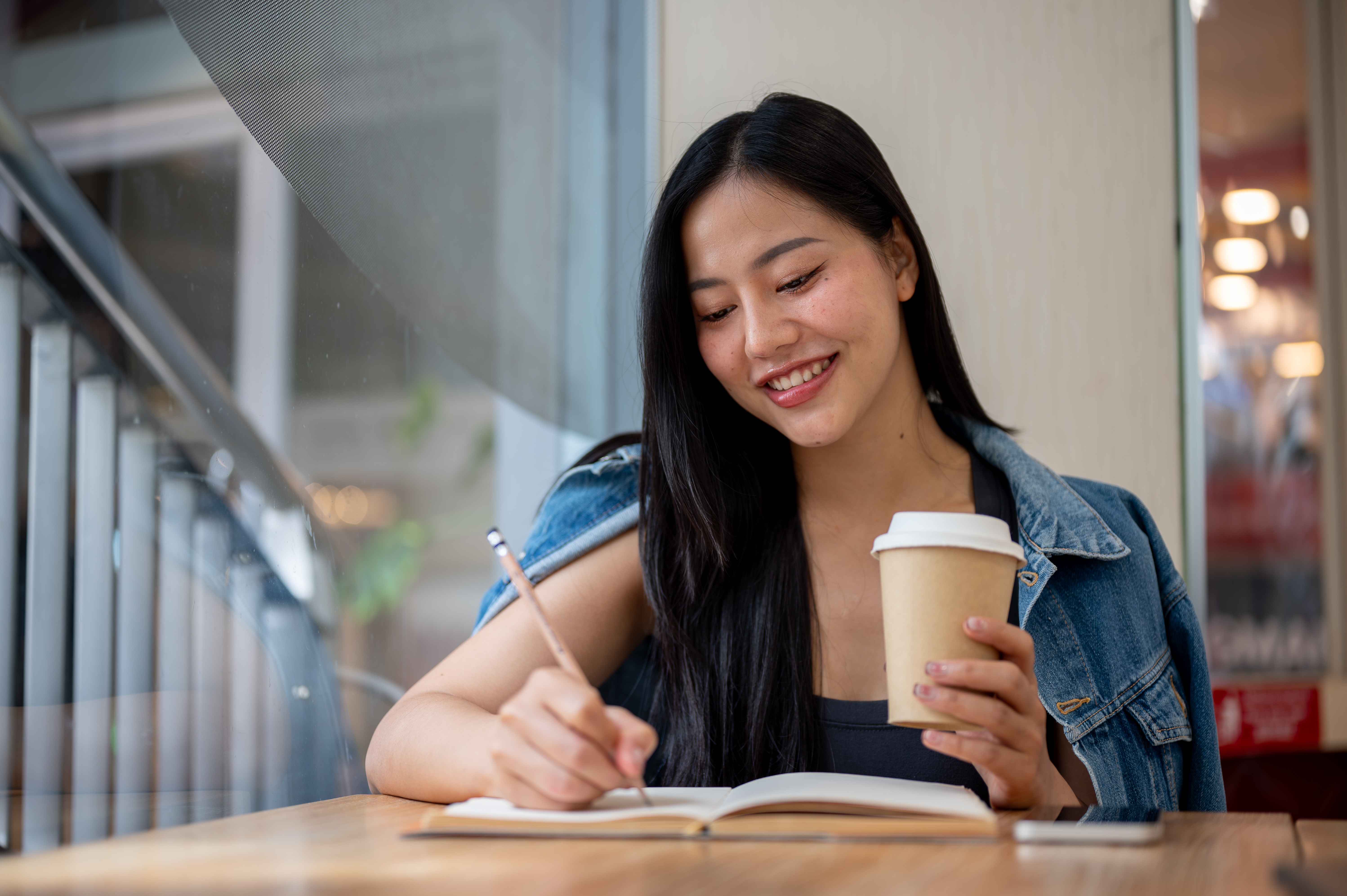 Person smiling while writing in a notebook and holding a coffee cup, seated at a cafe table. They&#x27;re wearing a denim jacket