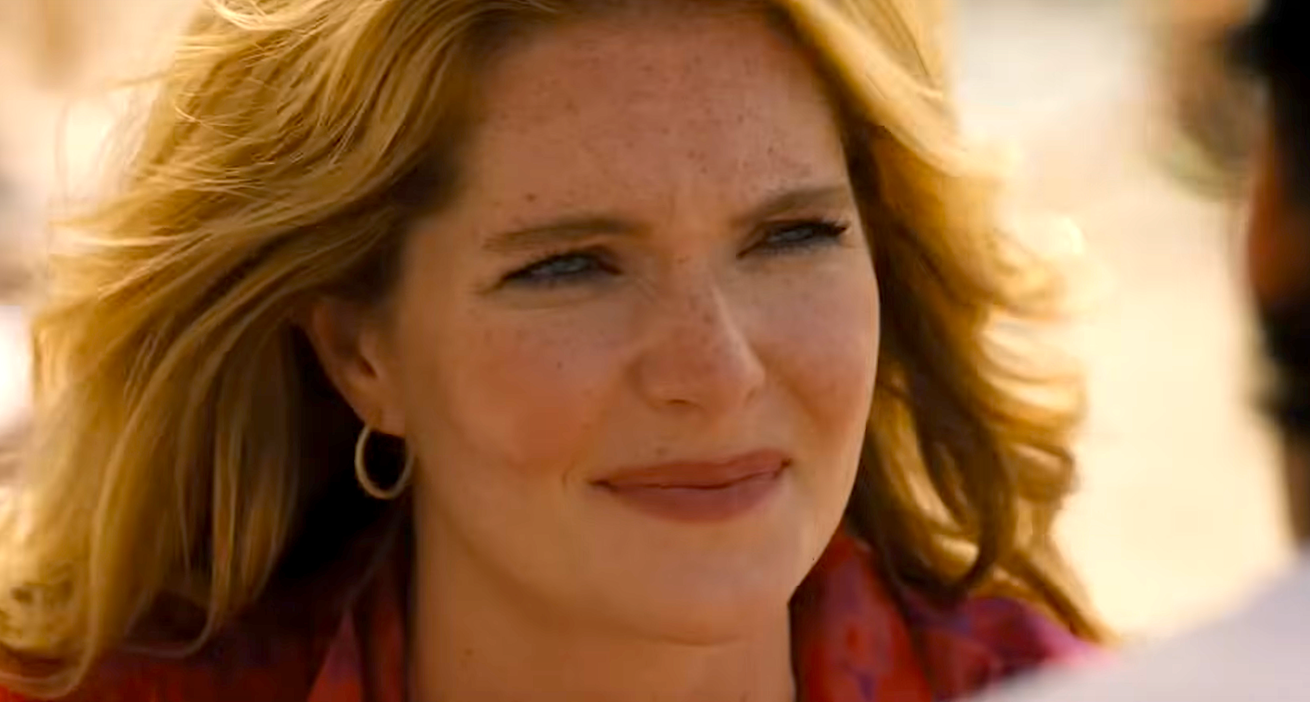 Person with wavy hair smiles slightly while looking at someone off-camera. Wearing hoop earrings and a patterned shirt