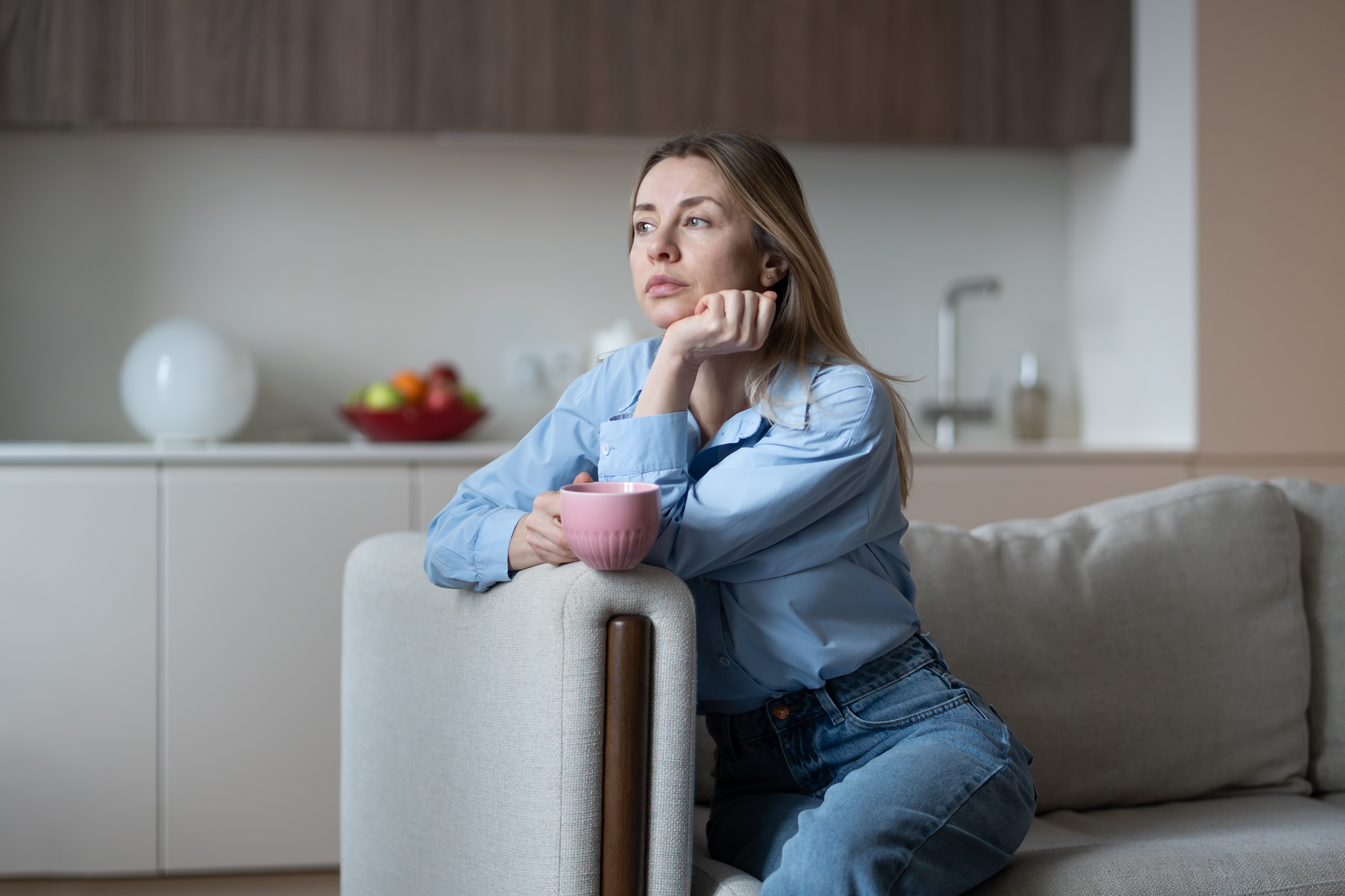 Person in a denim outfit sits on a couch, holding a cup, and gazes thoughtfully into the distance in a modern living room