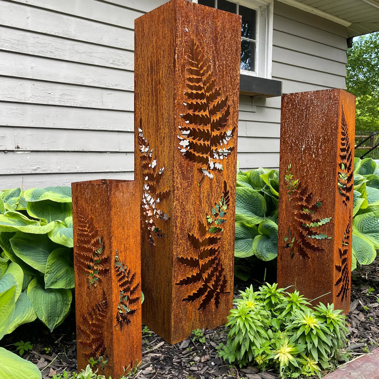 Three rust-colored metal garden sculptures with fern cutouts stand among green foliage
