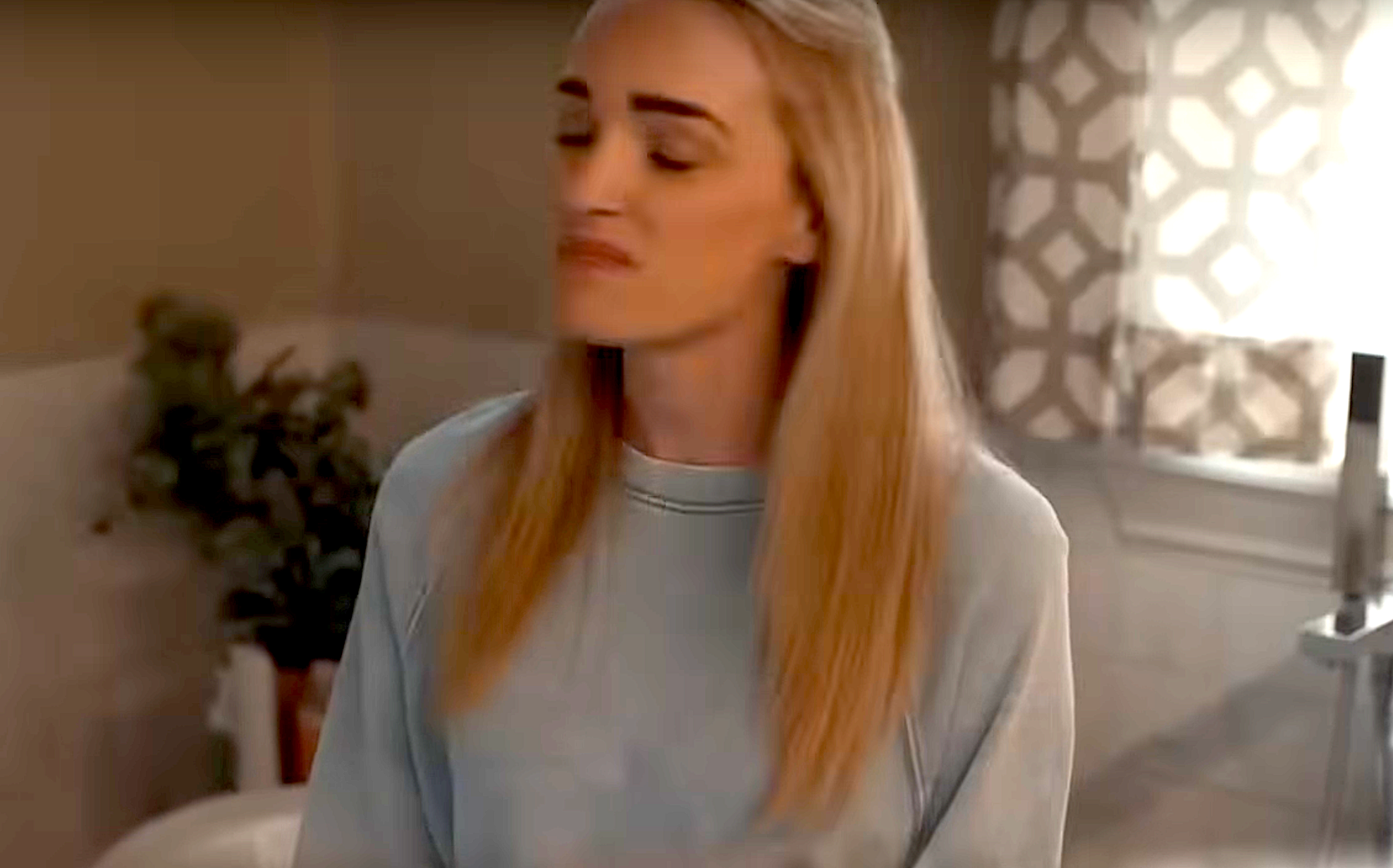 Person standing indoors, wearing a casual sweatshirt, with eyes closed and relaxed expression, beside a plant and patterned curtain