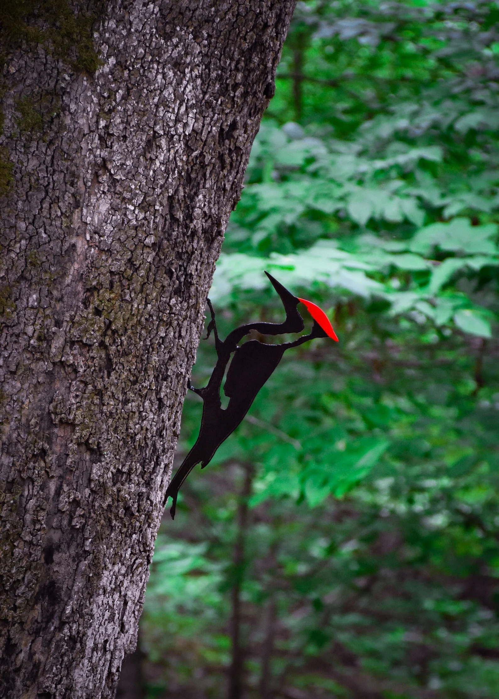 A metal woodpecker decoration clings to a tree trunk in a forest