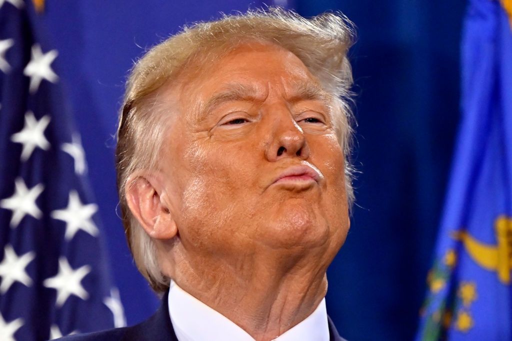 A person stands in front of a flag, making a pouting expression, with eyes slightly closed, head tilted back
