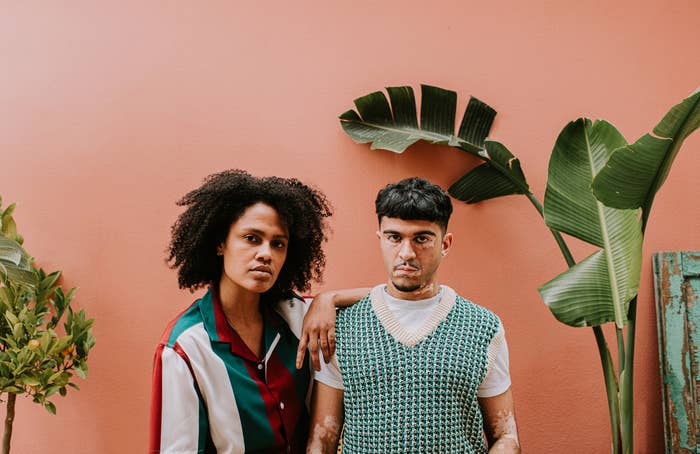 Two individuals stand against a solid background. One wears a chic, striped shirt, and the other sports a patterned vest. Green plants frame the composition