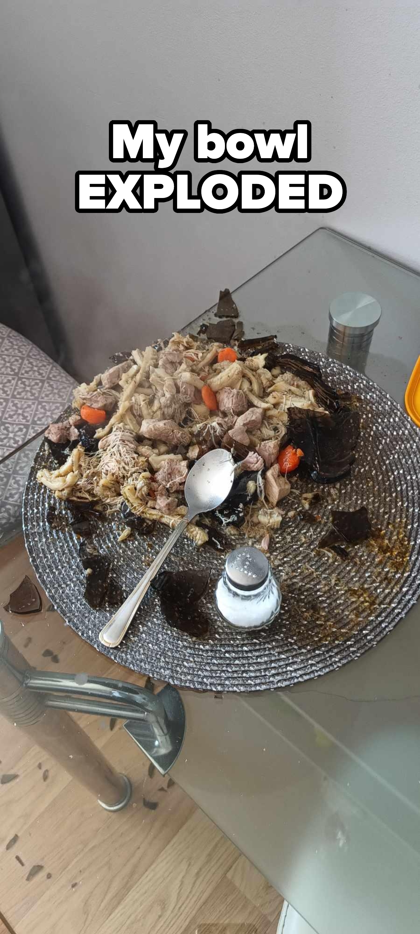 A broken glass plate on a table with scattered food, including pasta, meat, and vegetables, alongside a salt shaker and a napkin