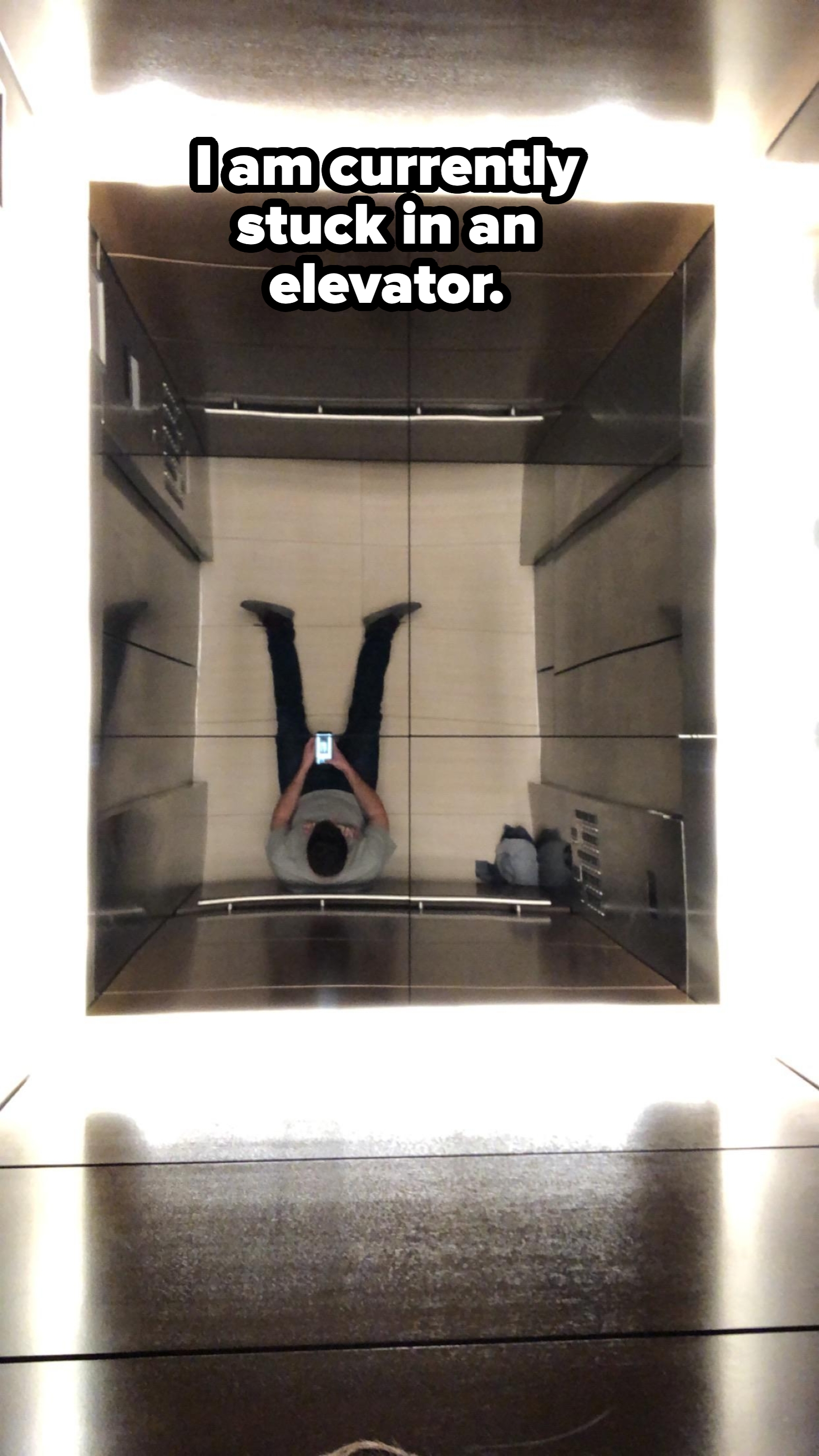 Person standing in an elevator taking a selfie in the mirrored ceiling, arms relaxed at sides, wearing casual attire