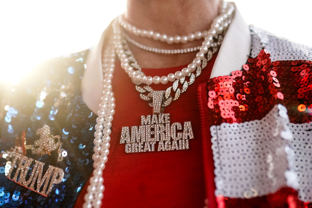 Person wearing a sequined jacket with a &quot;TRUMP&quot; patch and layered necklaces, including one with &quot;MAKE AMERICA GREAT AGAIN&quot; pendants