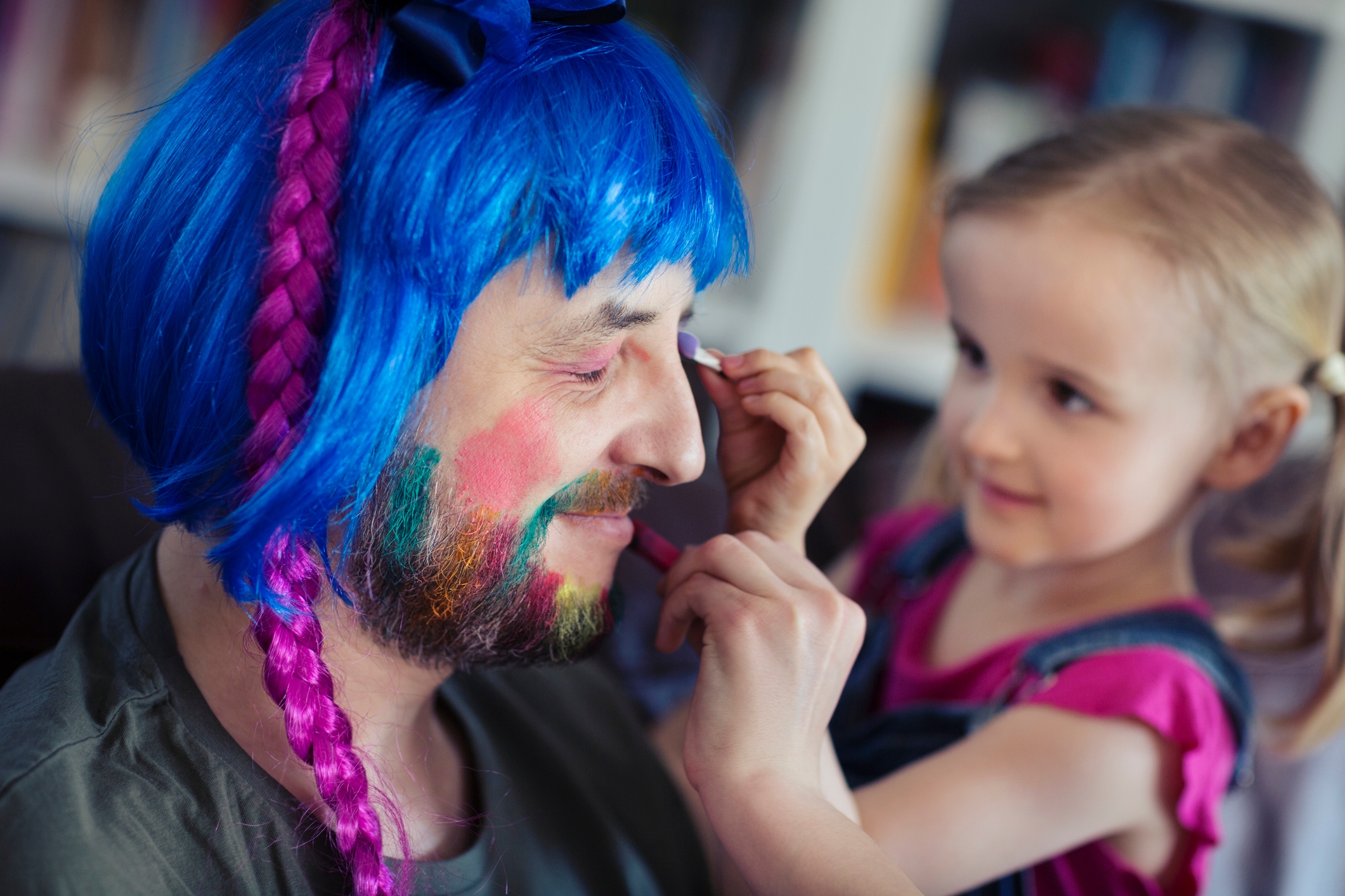 Child in pigtails playfully applies makeup to an adult wearing a blue wig with pink braids