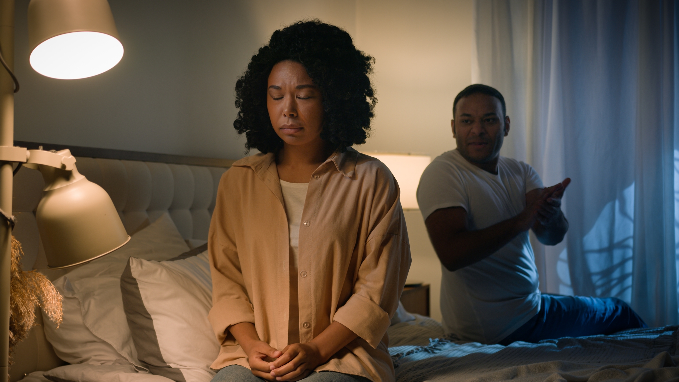 A person sits on a bed looking upset, while another sits behind them with a concerned expression. Both are in casual clothing