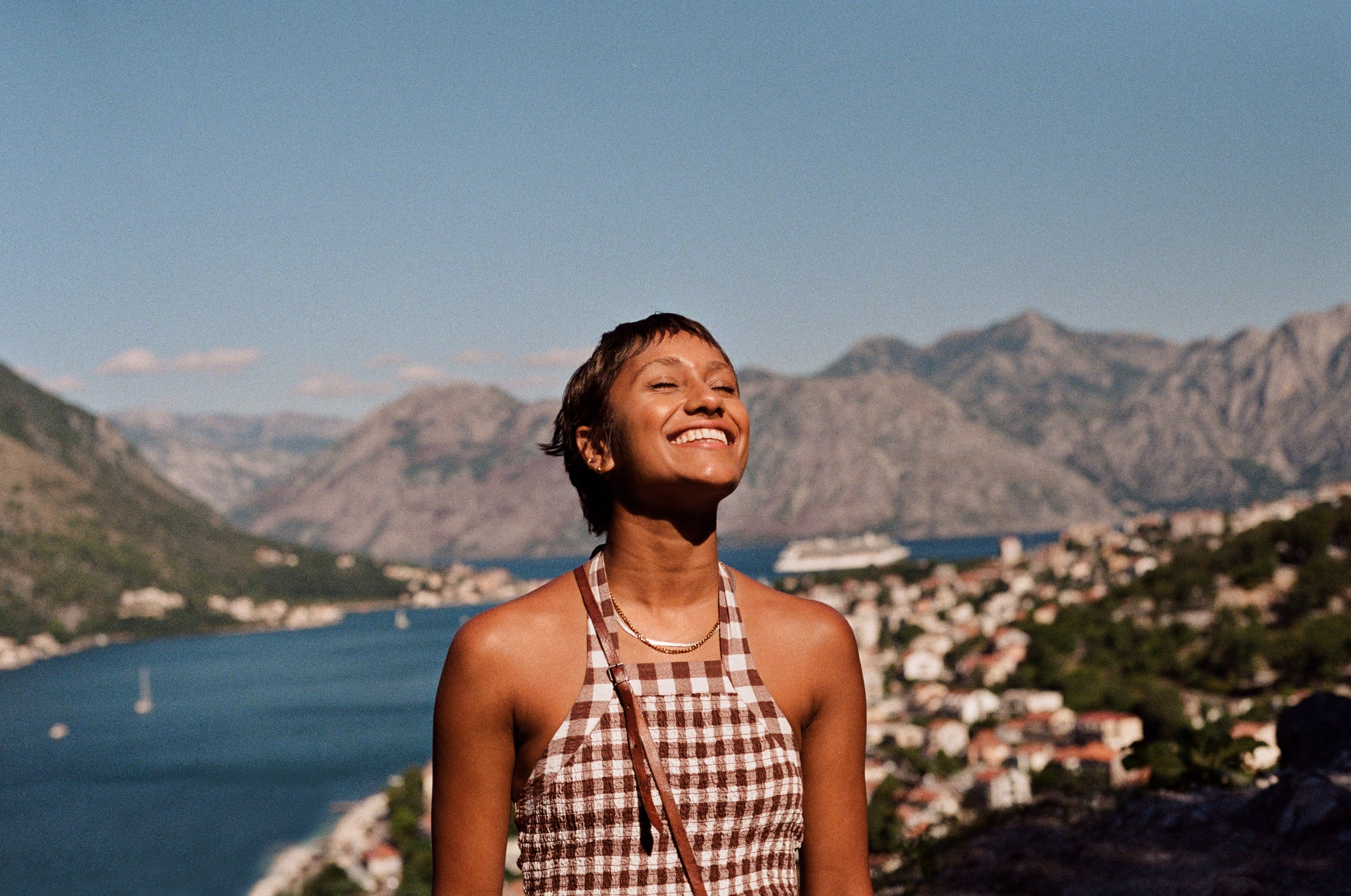 Person smiling joyfully with a scenic mountainous landscape in the background, featuring a winding river and a town on the banks