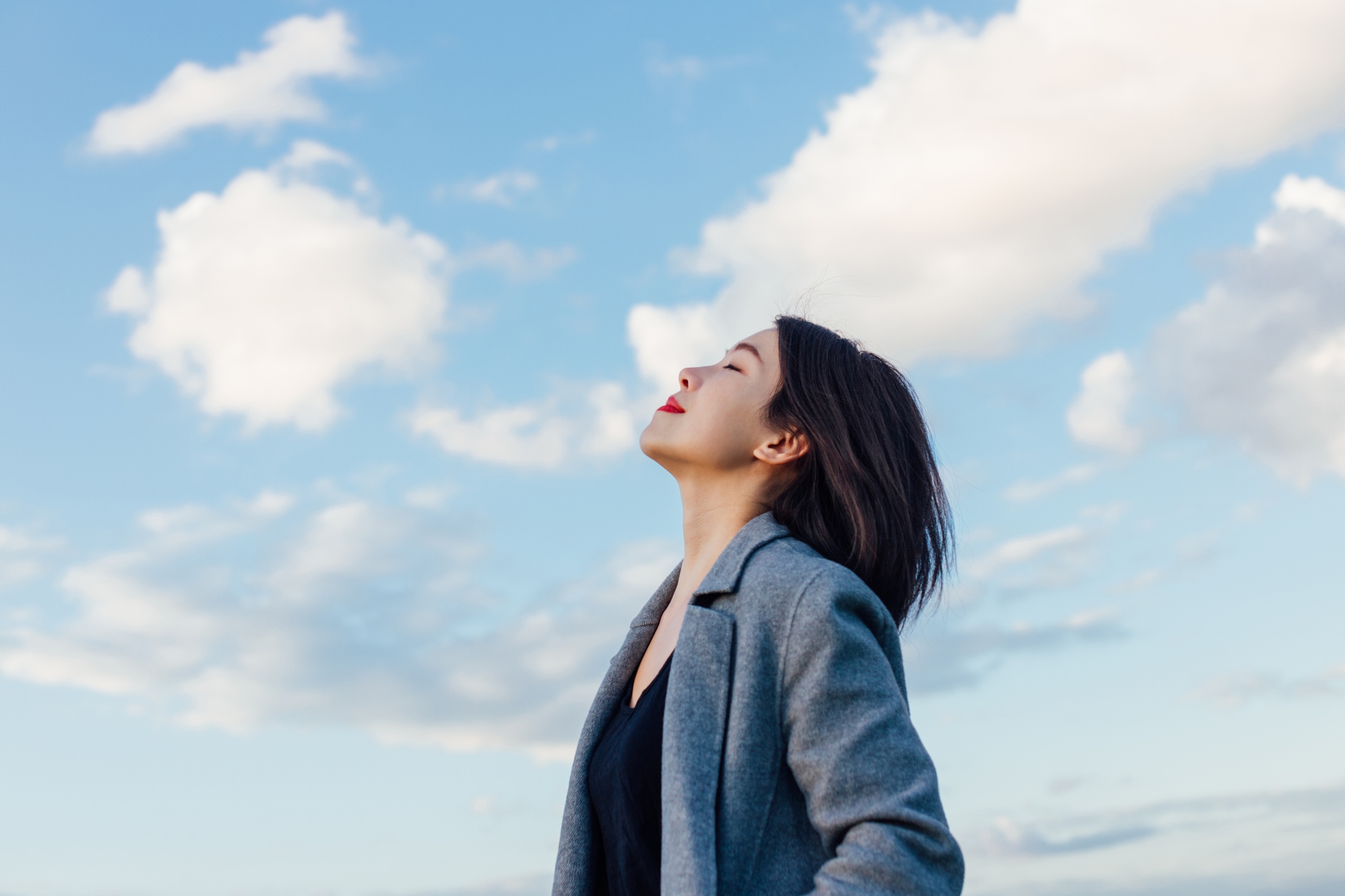 A woman with short hair in a coat stands outdoors, eyes closed, face tilted up, exuding a sense of tranquility against a cloudy sky