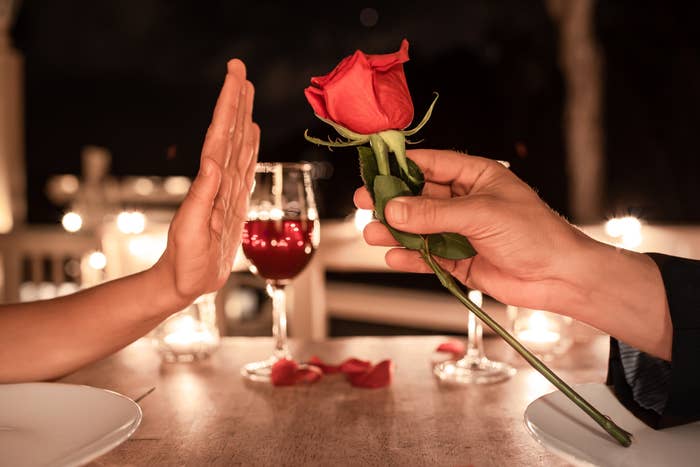 Person rejecting a red rose offered at a candlelit table, with wine glasses and rose petals visible