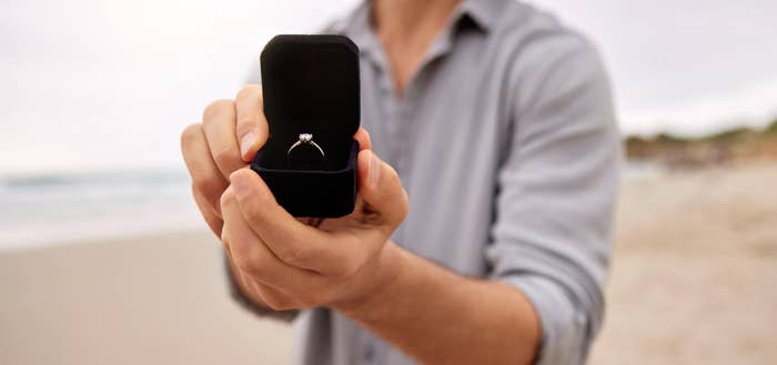 A person on a beach is holding an open ring box with a diamond engagement ring, offering it forward