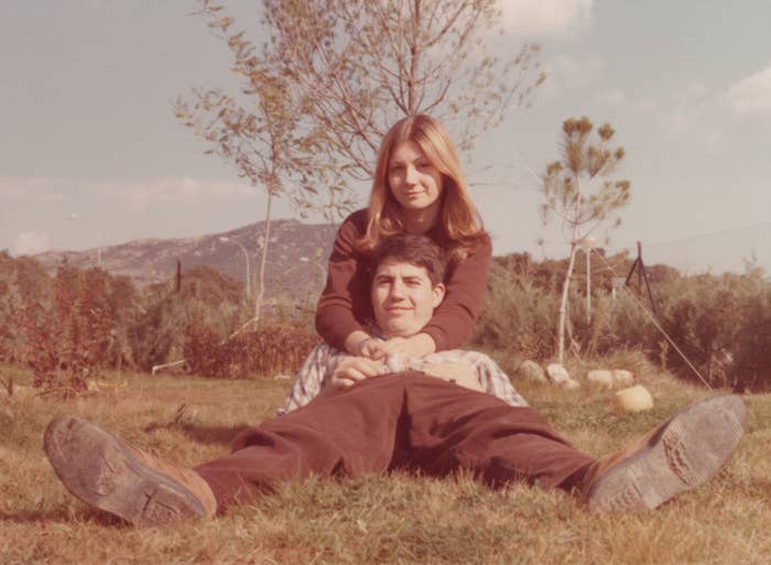 A young couple sits on grass, smiling warmly at the camera. The woman wraps her arms around the man, with scenic mountains in the background